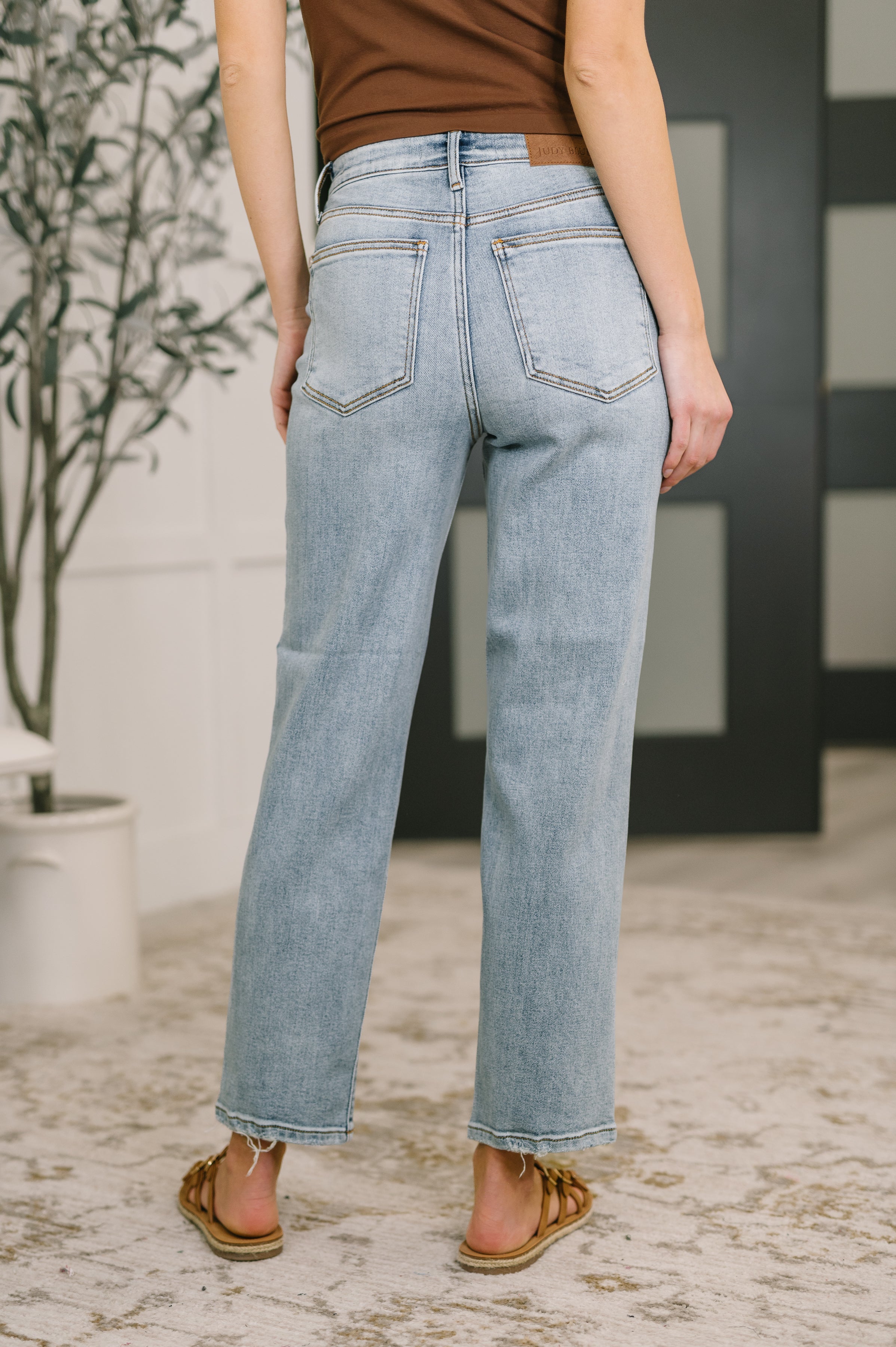 Back view of woman wearing light blue Natalie High Waist Ankle Straight Jeans with raw hem and sandals indoors