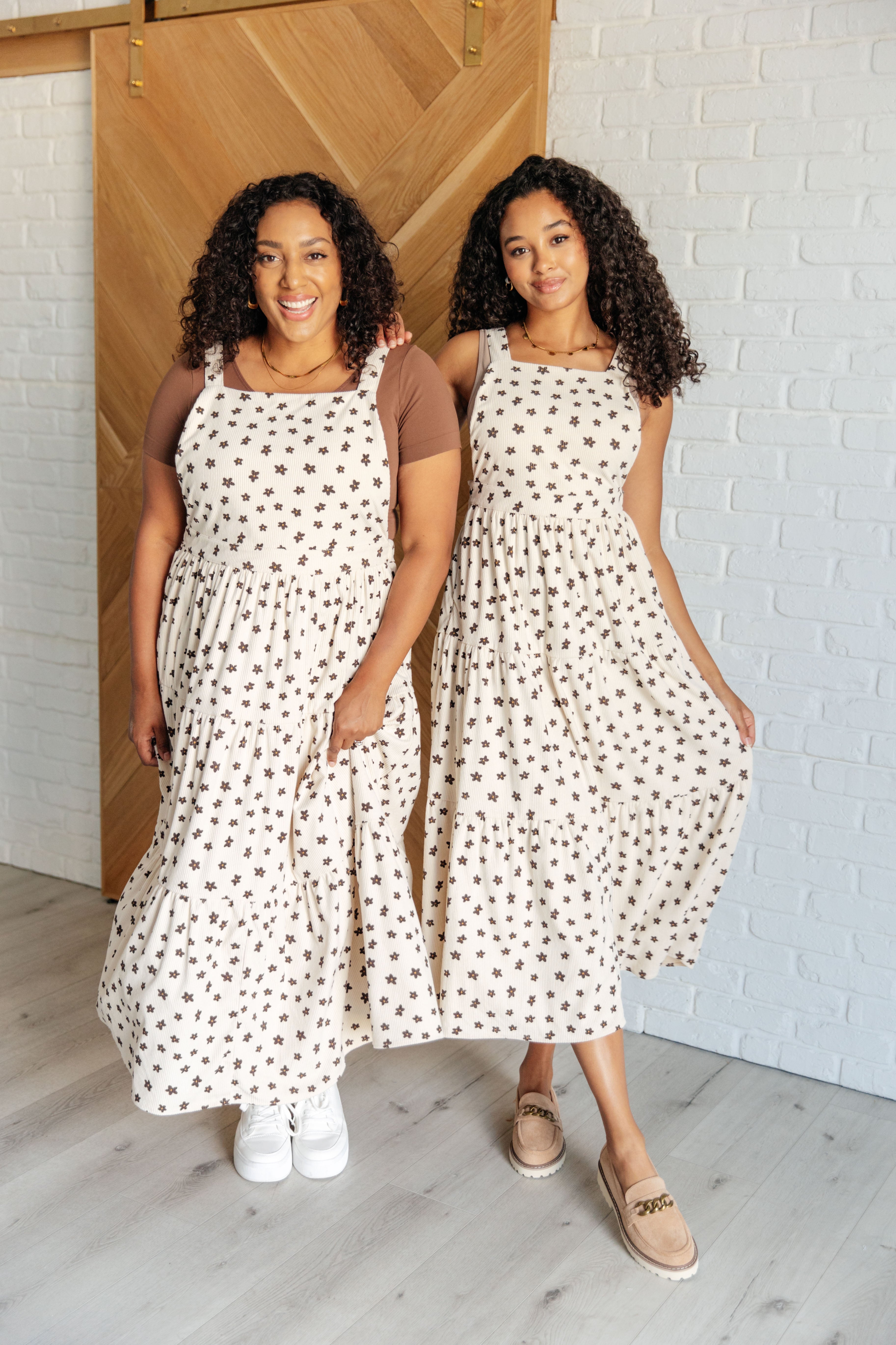Two women wearing cream sleeveless dresses with brown floral prints posing indoors by a wooden door and white brick wall