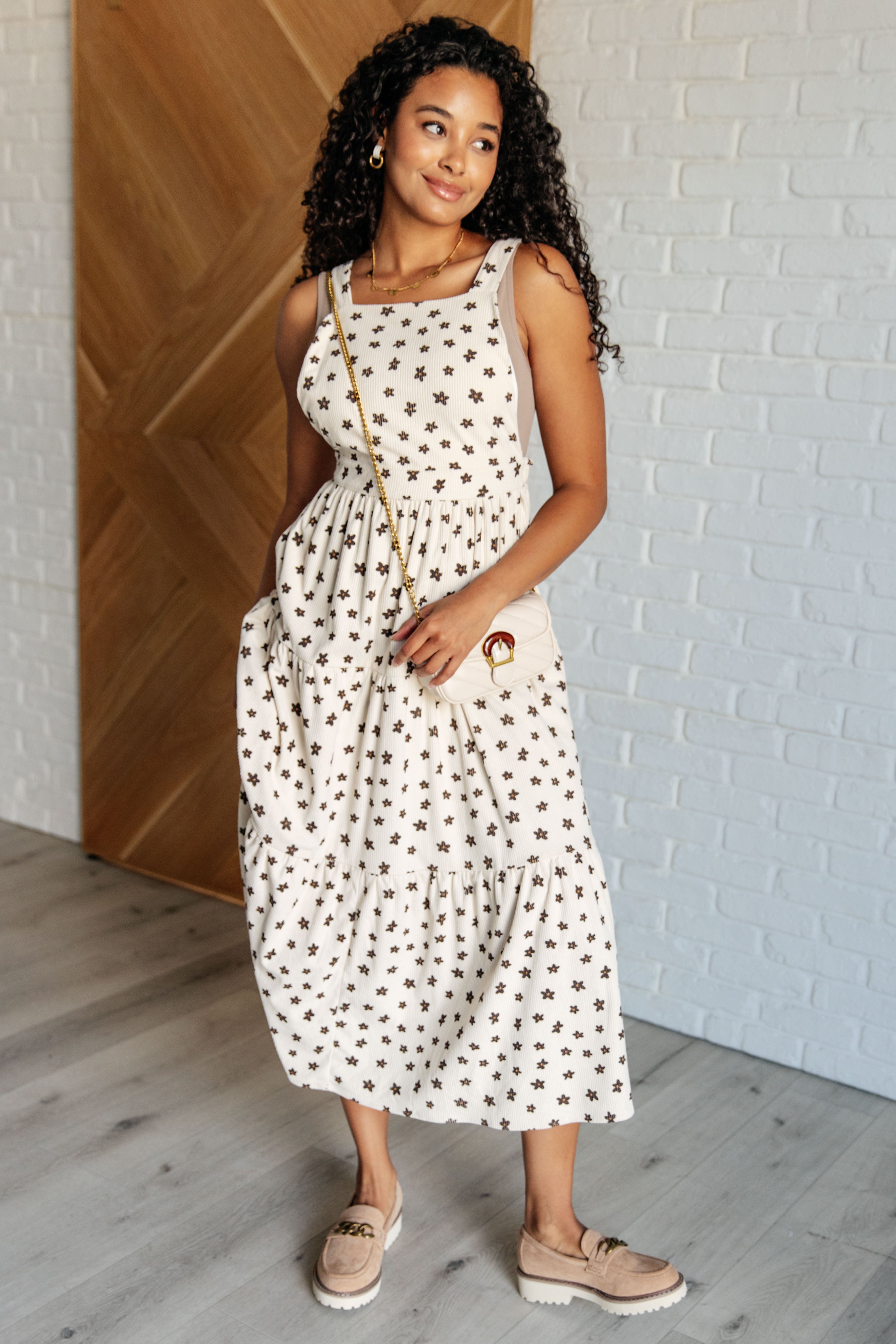 Woman wearing a cream sleeveless dress with dark star patterns and beige shoes standing indoors by a wooden door and white brick wall