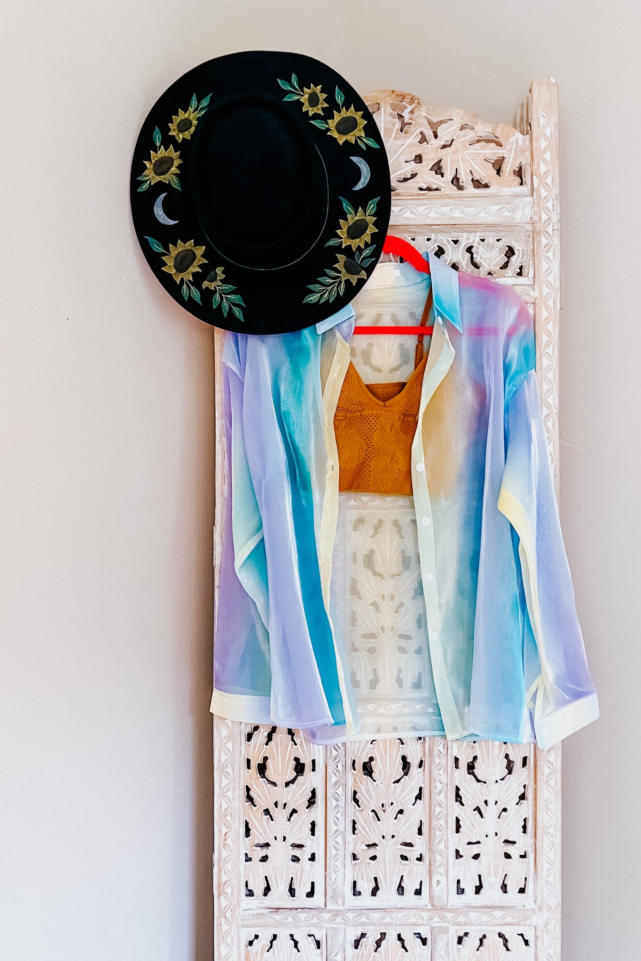 Sheer button-up top in soft blue and pastel hues layered over a mustard knit crop top, displayed on a carved white wooden panel with a black sunflower and moon embroidered hat hanging above.