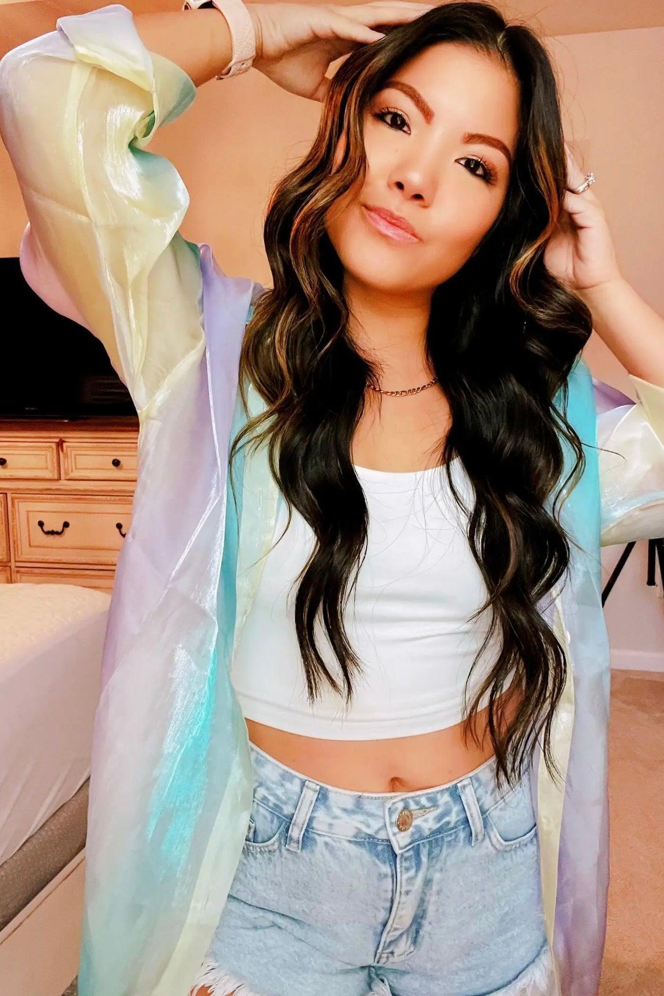 Young woman wearing a pastel blue and yellow sheer iridescent button-up top over a white crop tank with high-rise light denim shorts indoors