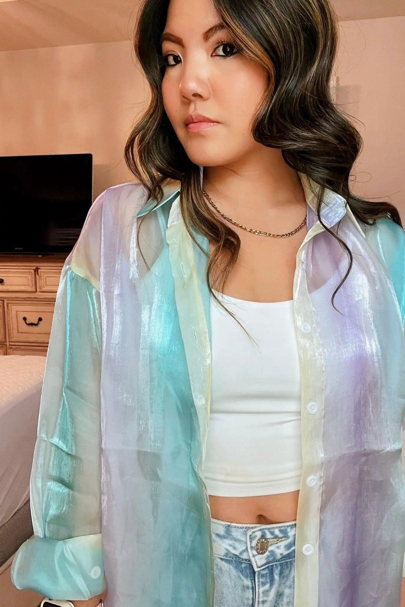 Woman indoors wearing a sheer, blue iridescent button-up top layered over a white crop tank and light-wash jeans with loose waves in her hair.