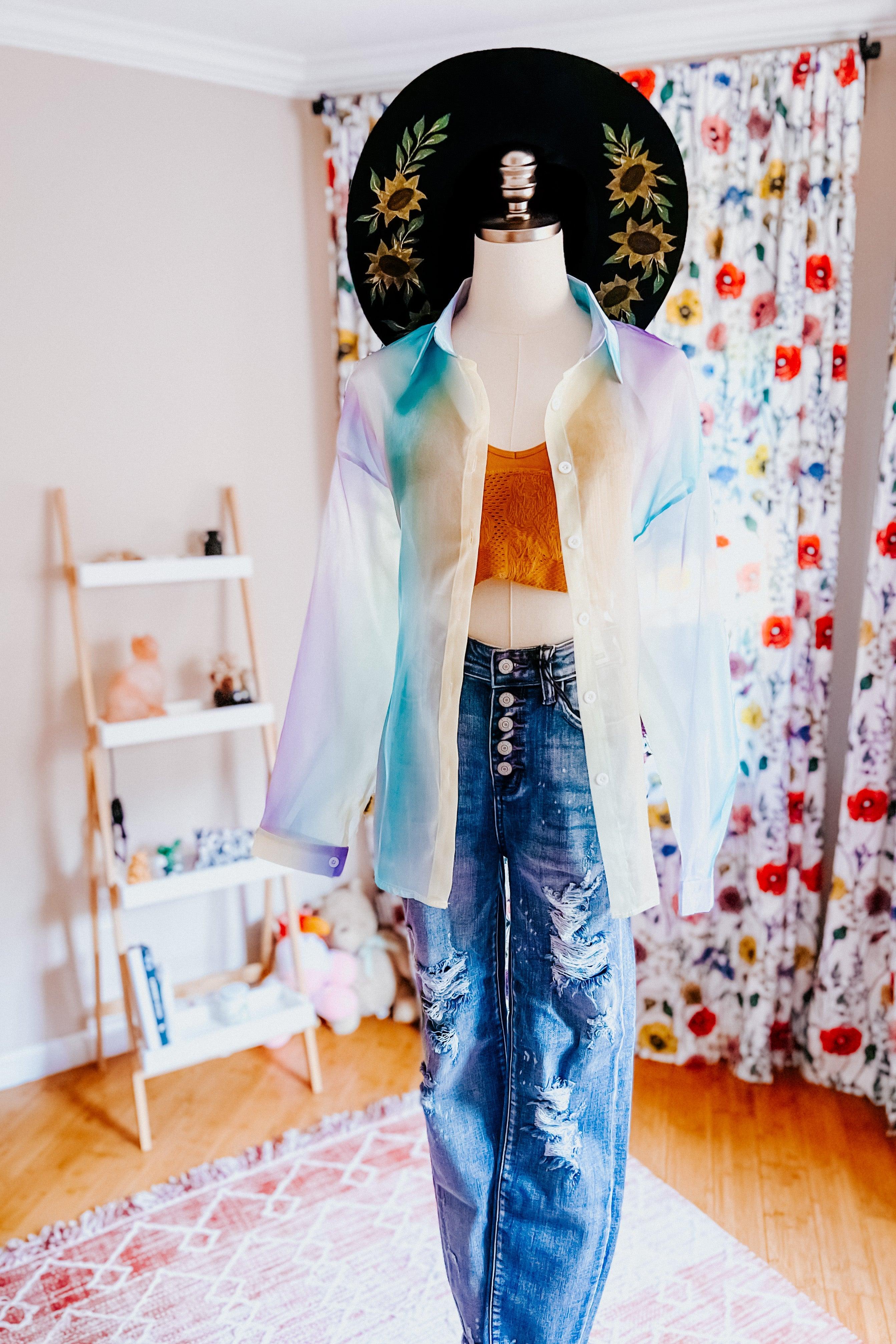 Mannequin dressed in a sheer, pastel blue iridescent button-up top layered over a mustard crop top with distressed high-waisted jeans and a black wide-brimmed hat with floral embroidery in a cozy room.