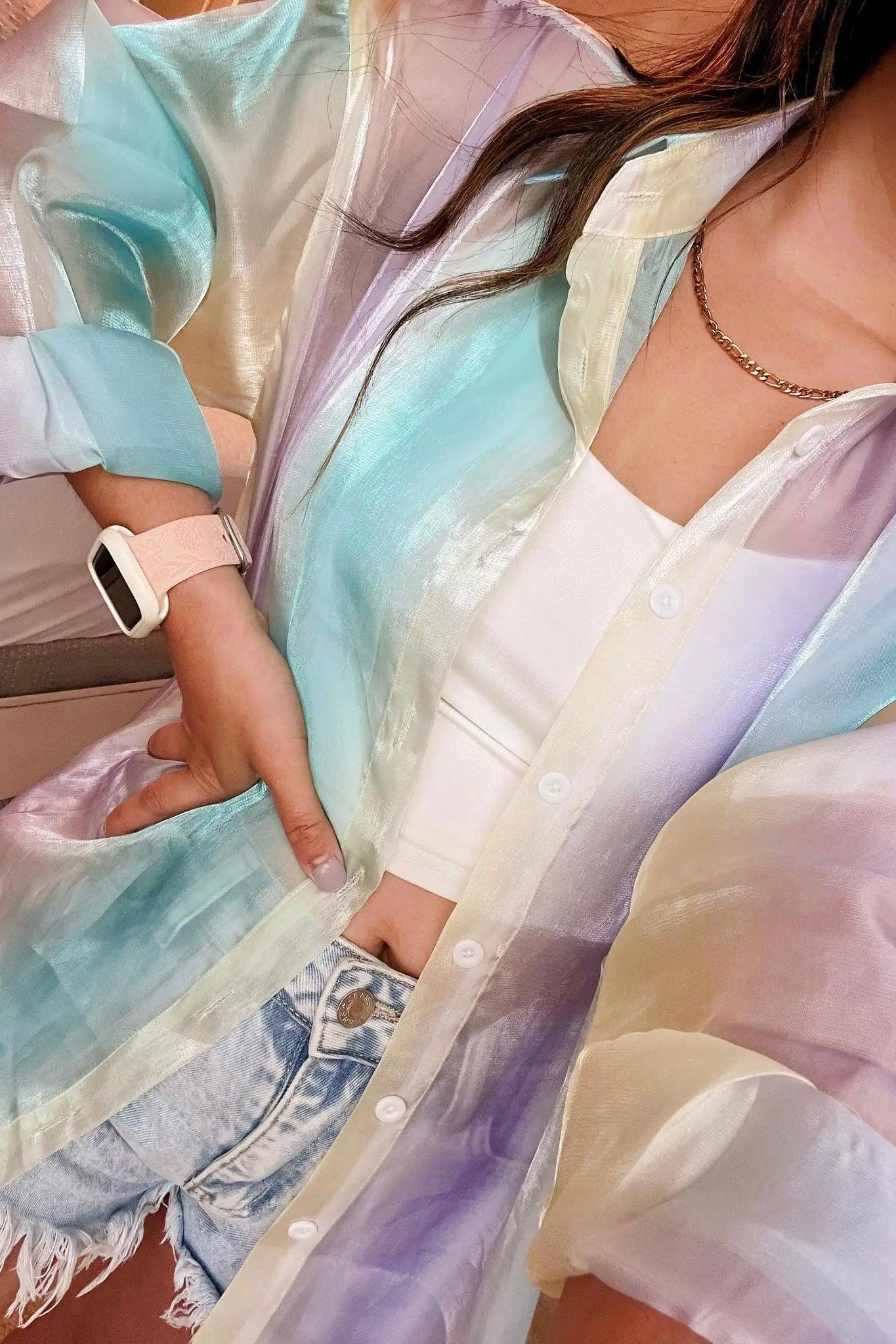 Close-up of person wearing a sheer, pastel blue and lavender iridescent button-up top layered over a white crop top and light-wash denim shorts with a chain necklace and smartwatch.