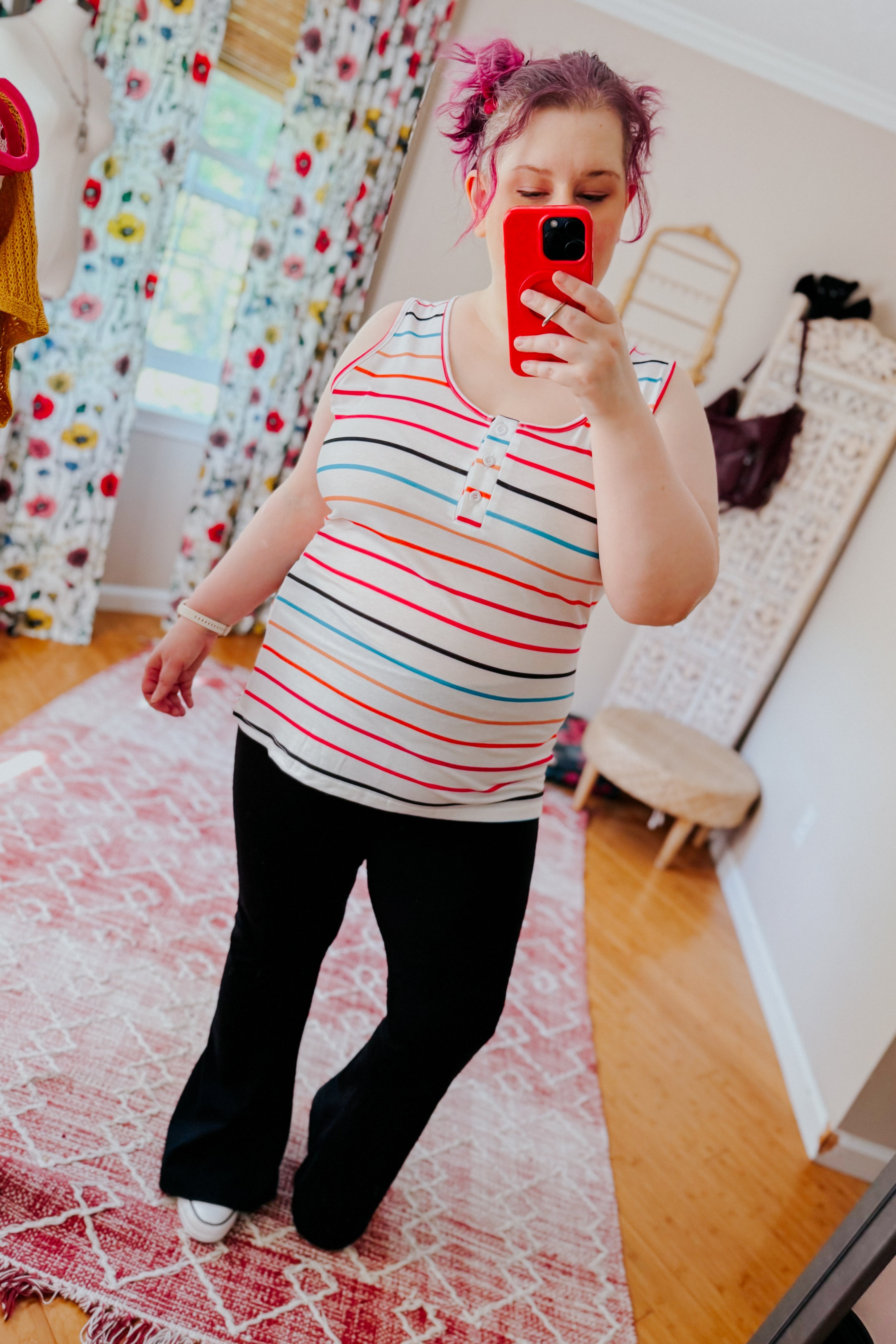 Woman with purple hair taking mirror selfie wearing ivory henley tank top with colorful stripes and black pants indoors