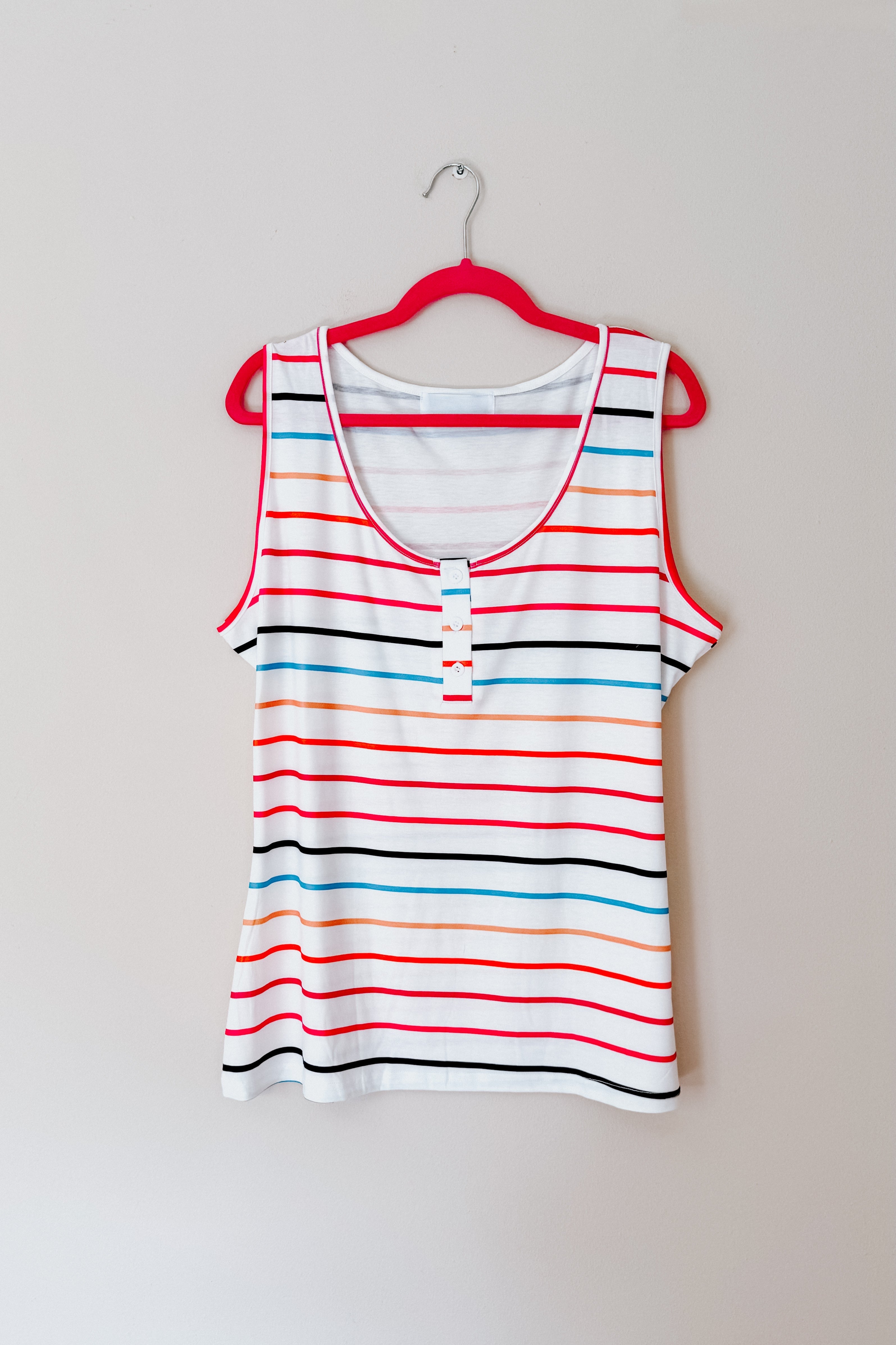 Ivory sleeveless Henley tank with colorful horizontal stripes and red trim hanging on pink hanger against white wall