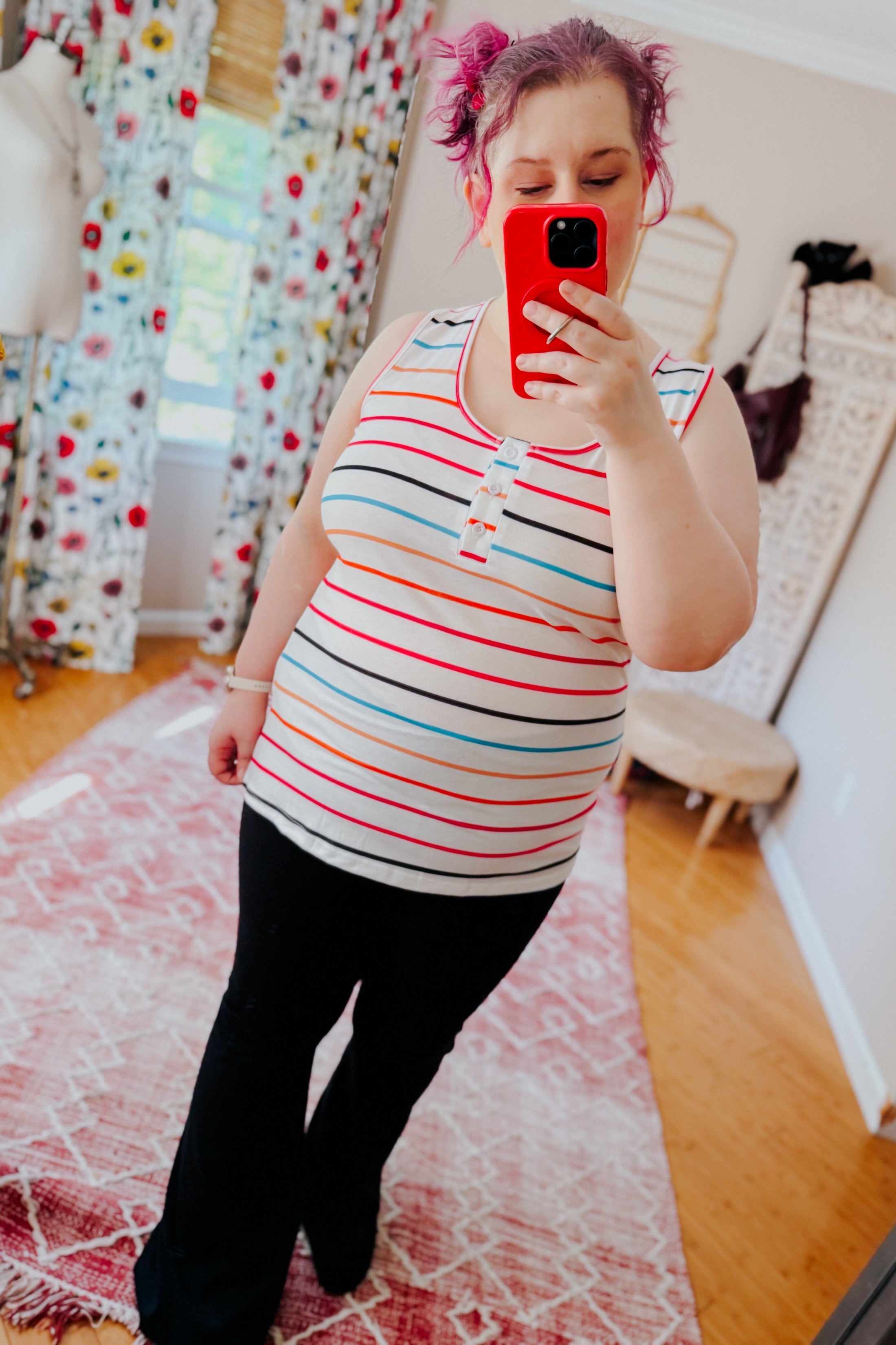 Woman wearing ivory sleeveless henley tank with colorful stripes and black pants taking mirror selfie indoors