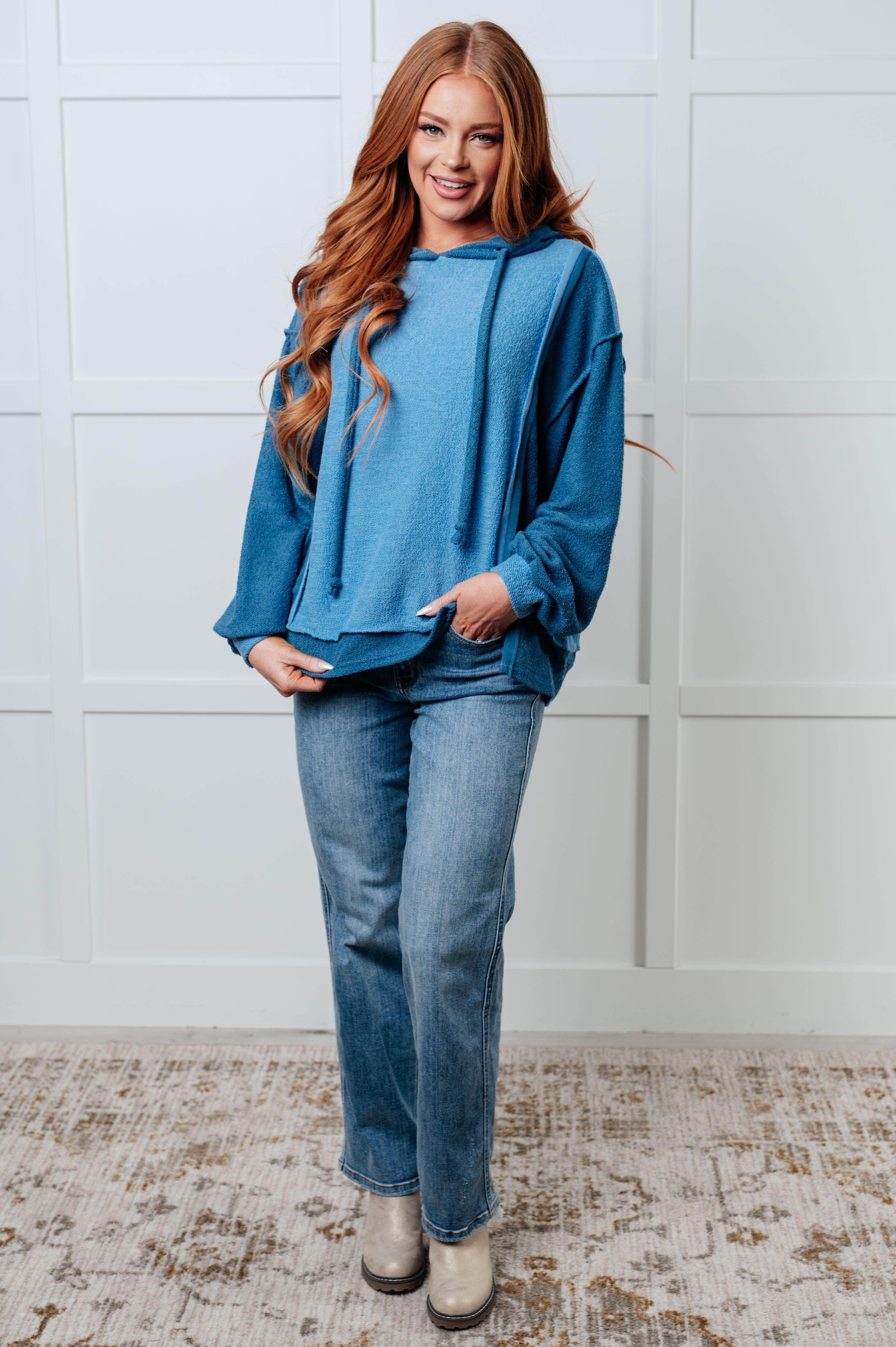 Woman indoors wearing a relaxed fit blue color block hoodie with long sleeves paired with straight-leg light wash jeans and beige ankle boots smiling