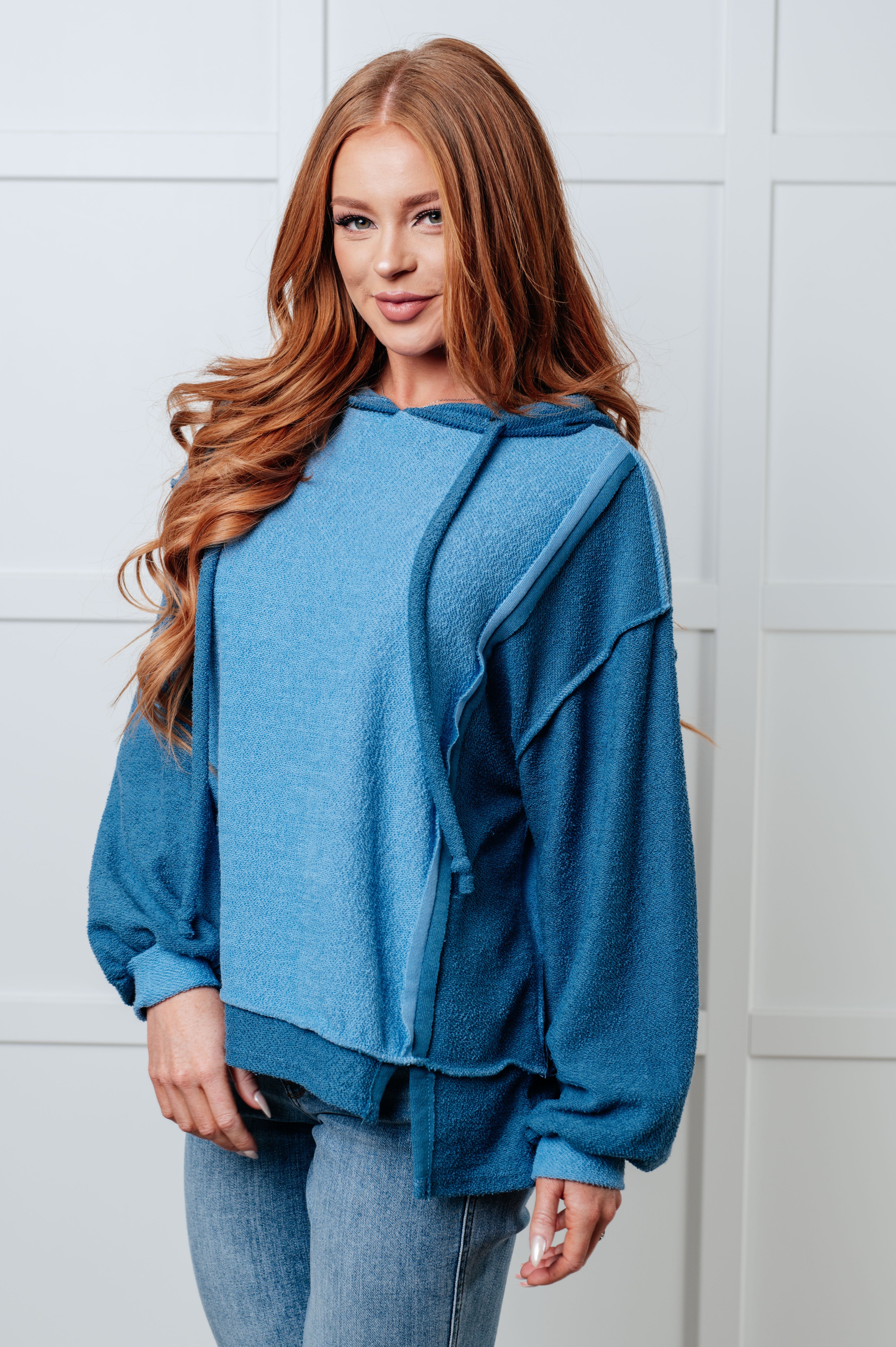 Woman with long red hair wearing a relaxed fit blue color block hoodie with a textured fabric and light wash jeans, standing indoors.