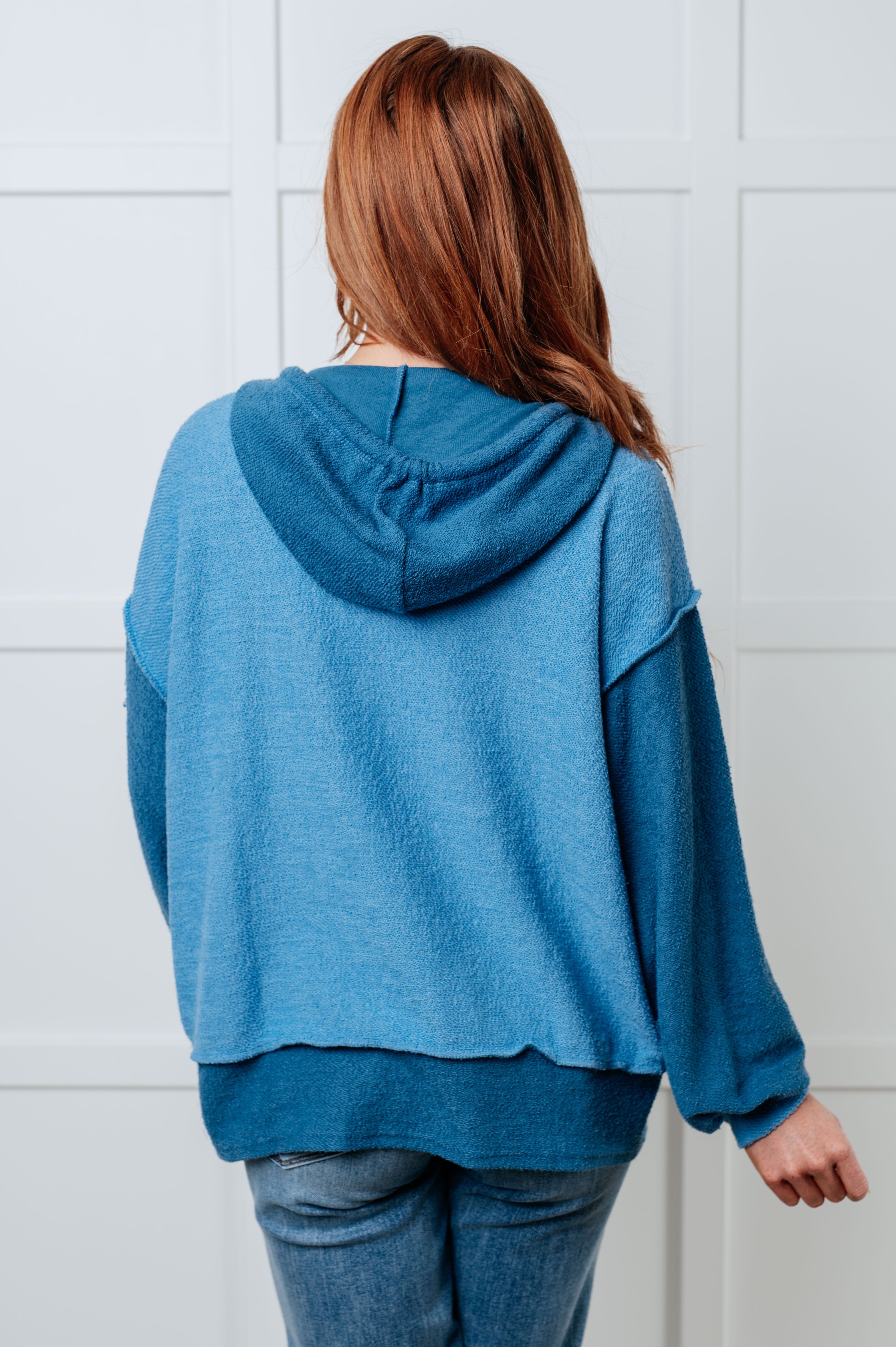 Woman wearing a relaxed fit blue color block hoodie with a textured fabric and hood, paired with light wash jeans, shown from behind indoors.