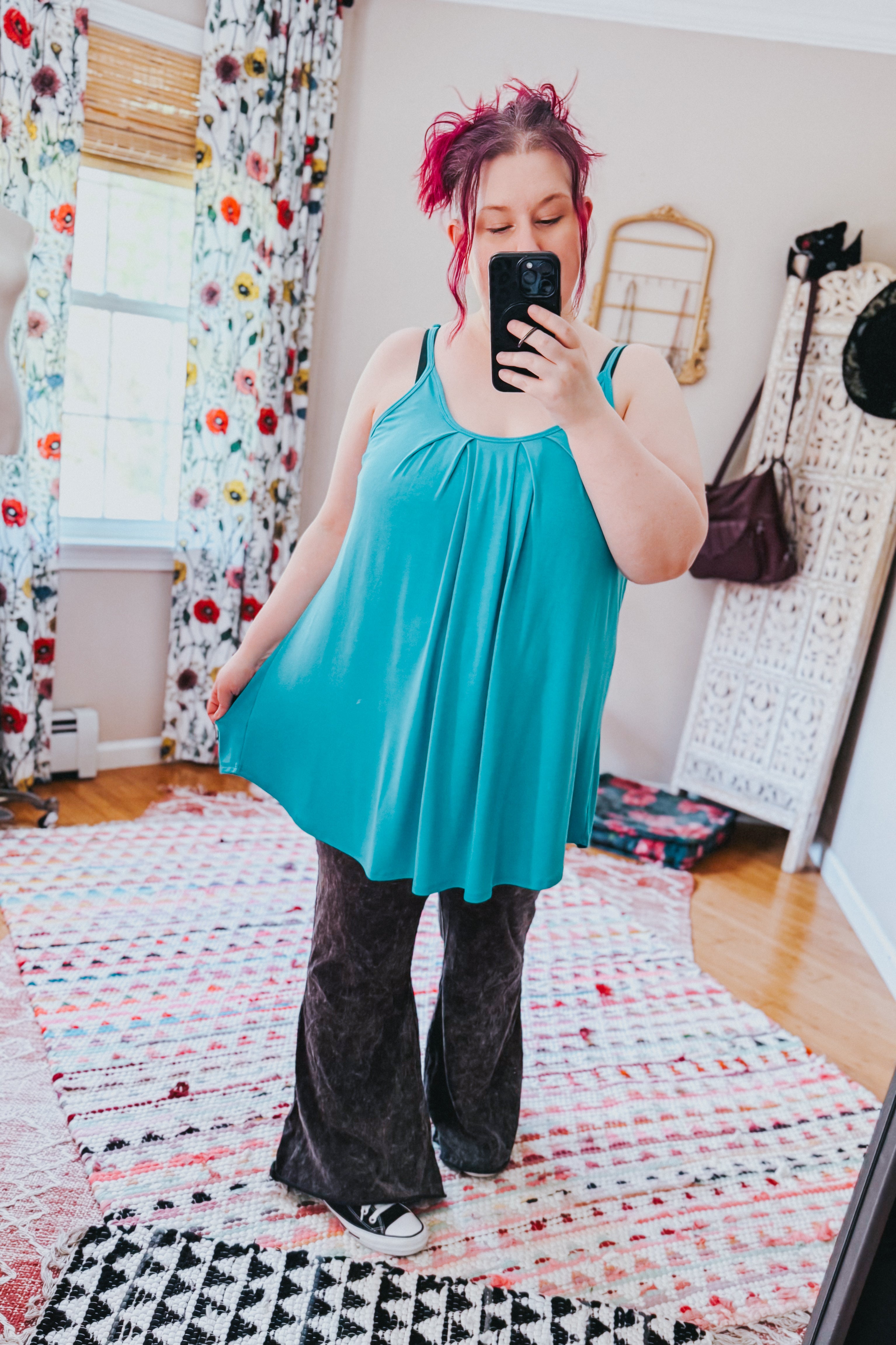 Woman taking a mirror selfie wearing a loose neon blue pleated tank top with thin straps paired with dark flared pants in a bright room with floral curtains and patterned rugs.