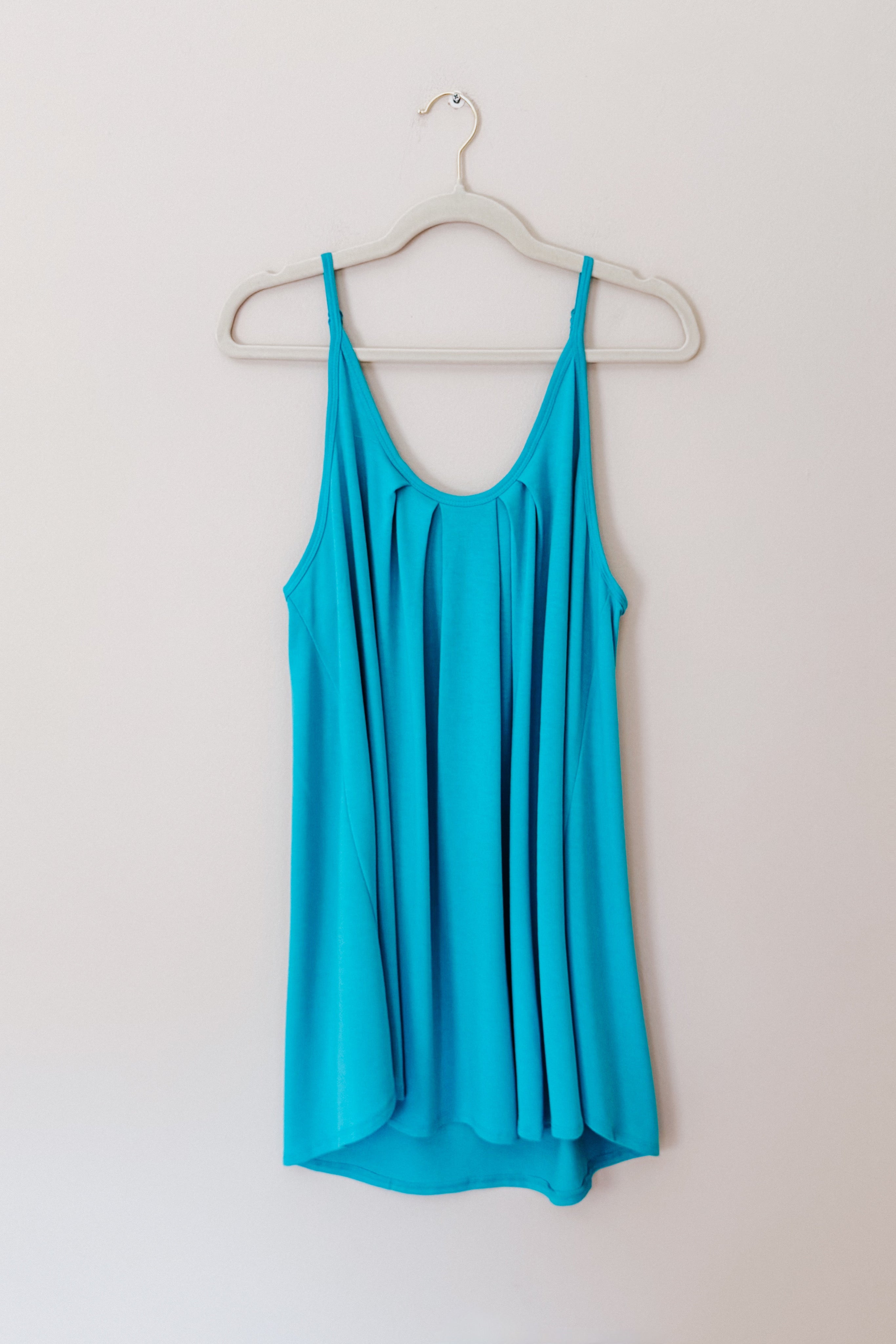 Bright neon blue pleated tank top with thin straps hanging on a white hanger against a plain light wall background.