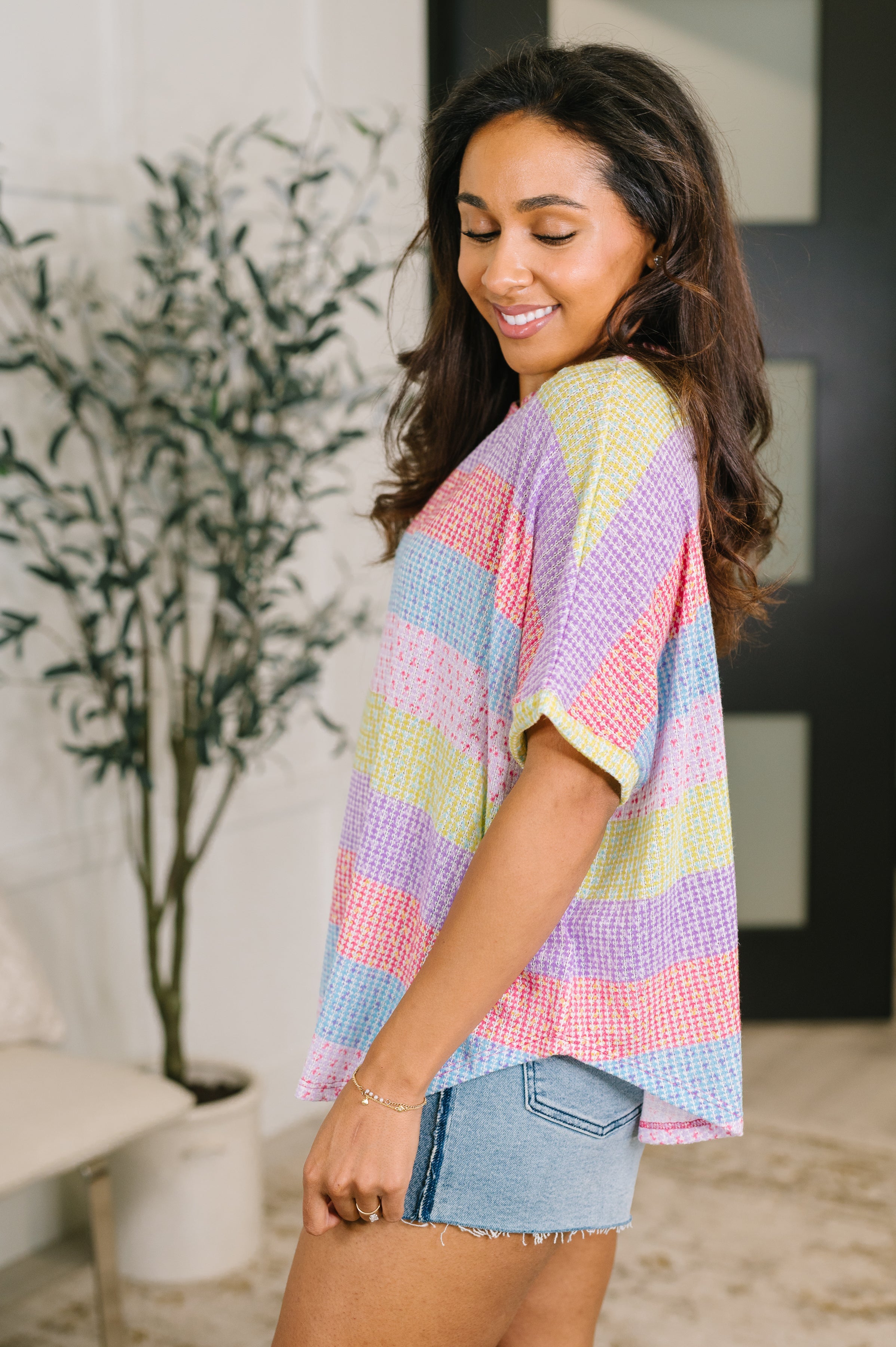 Side profile of woman smiling wearing relaxed pastel striped textured knit top with dolman sleeves and denim shorts indoors