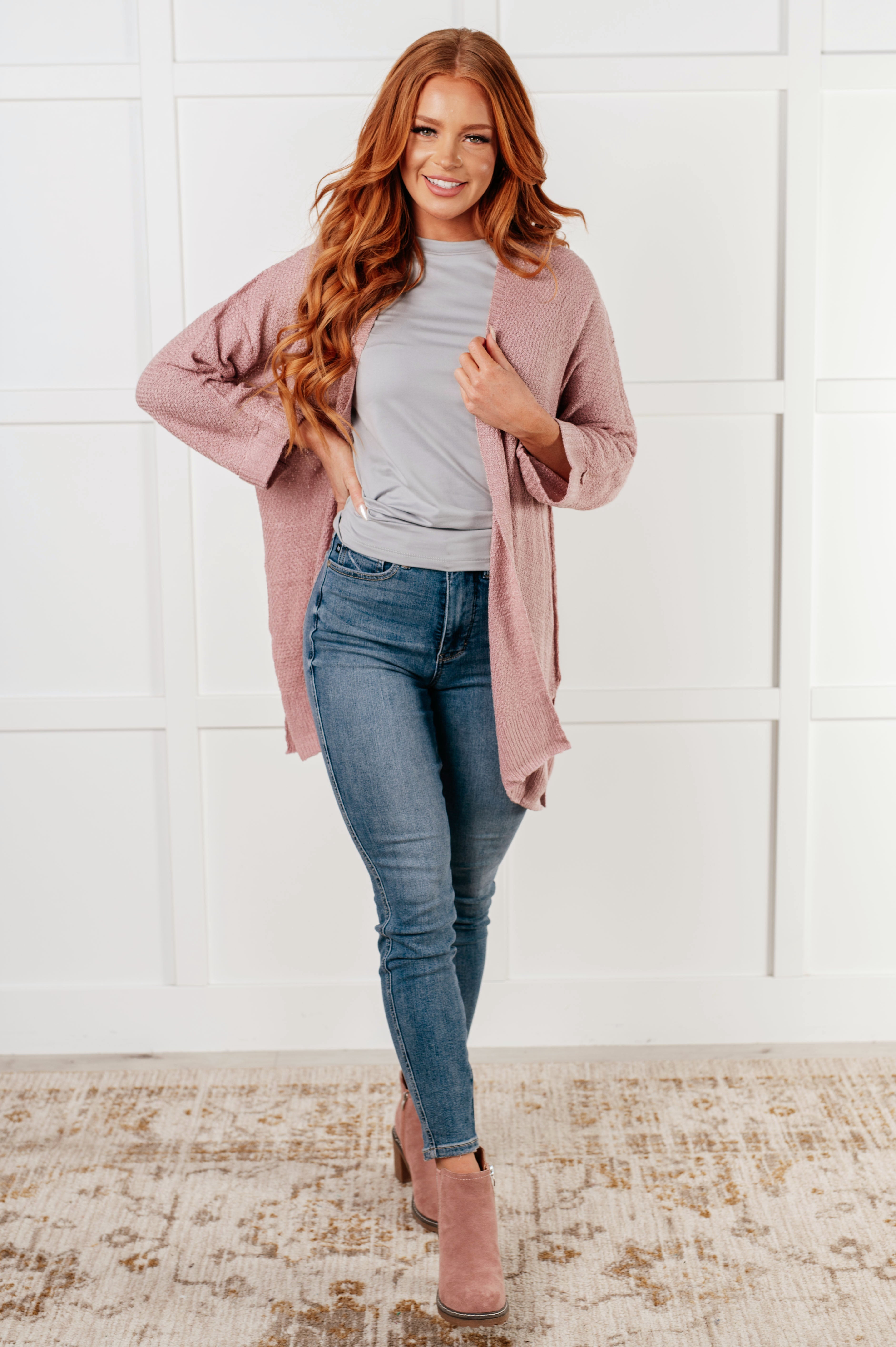 Woman posing indoors wearing a soft pink open-front cardigan layered over a light gray top and paired with slim blue jeans and pink ankle boots.
