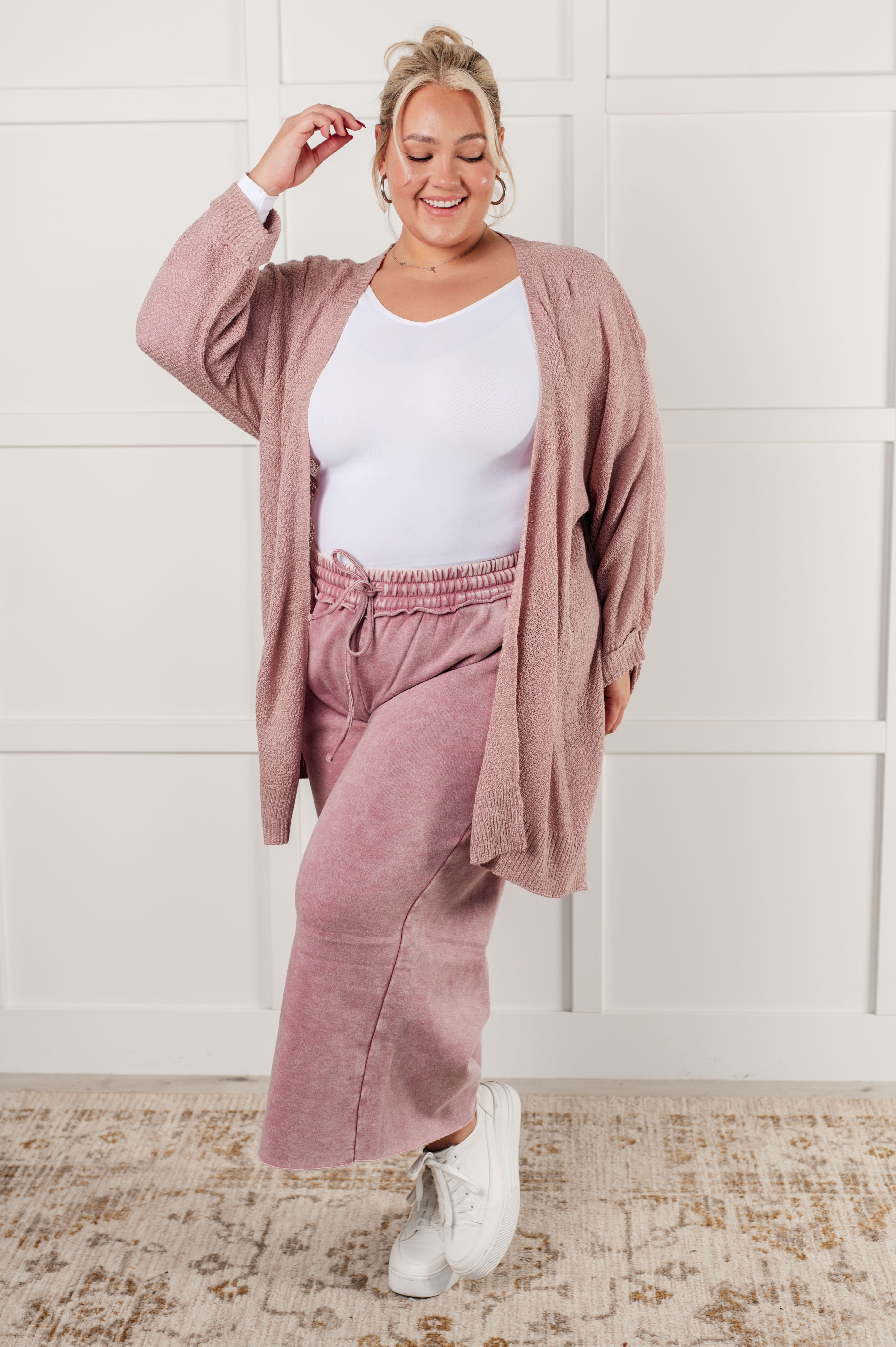 Smiling woman wearing a light pink open-front cardigan layered over a white fitted top and matching pink wide-leg pants with white sneakers, standing indoors.