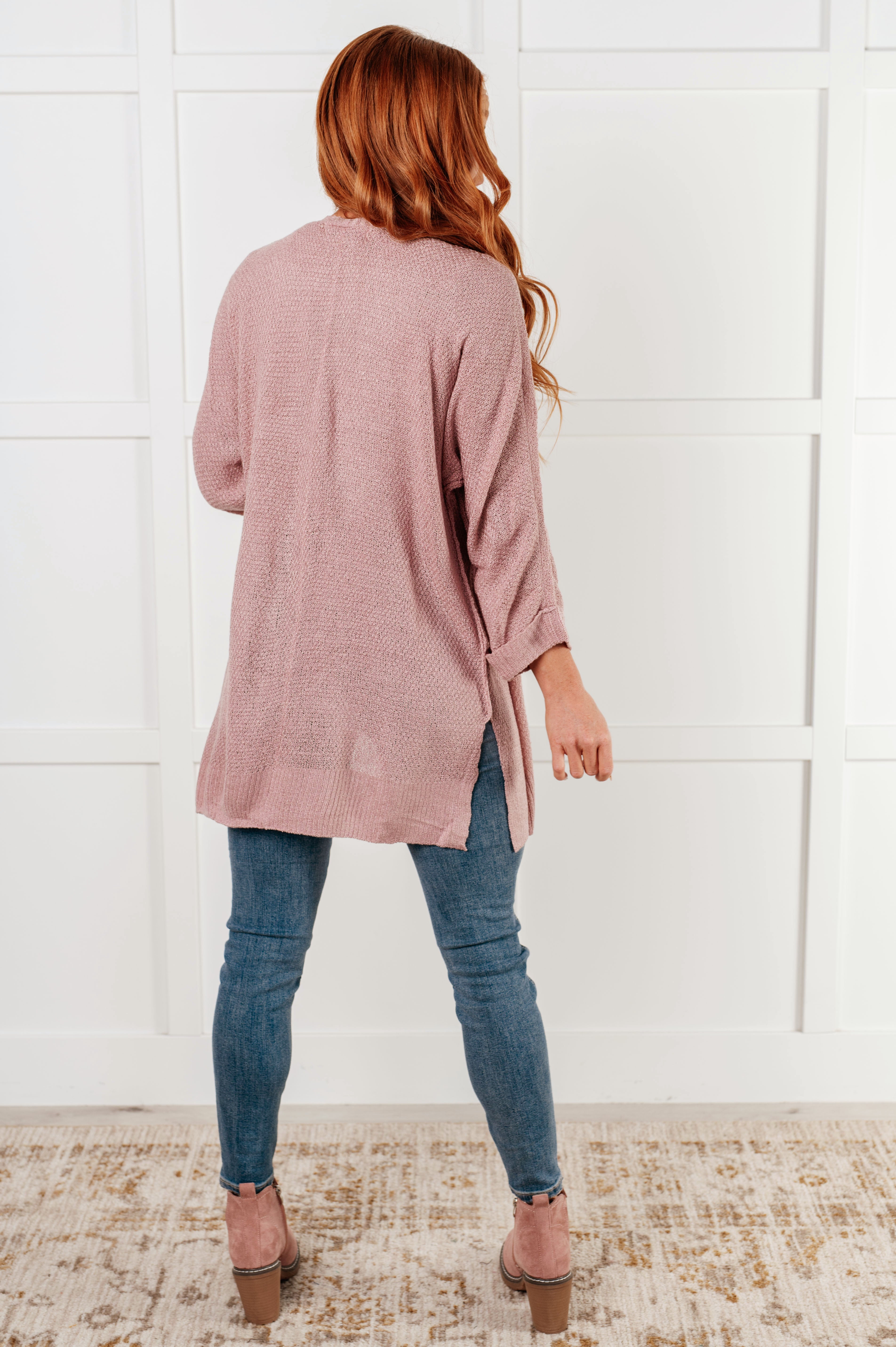 Woman standing indoors wearing a soft pink cardigan with rolled sleeves paired with fitted blue jeans and pink suede ankle boots.