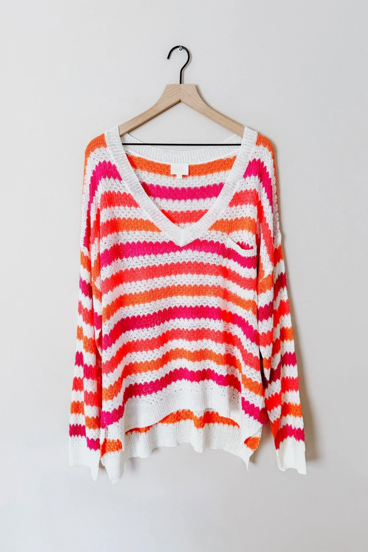 Light knit sweater with bold pink, orange, and white horizontal stripes and a relaxed fit hanging on a wooden hanger against a plain wall.