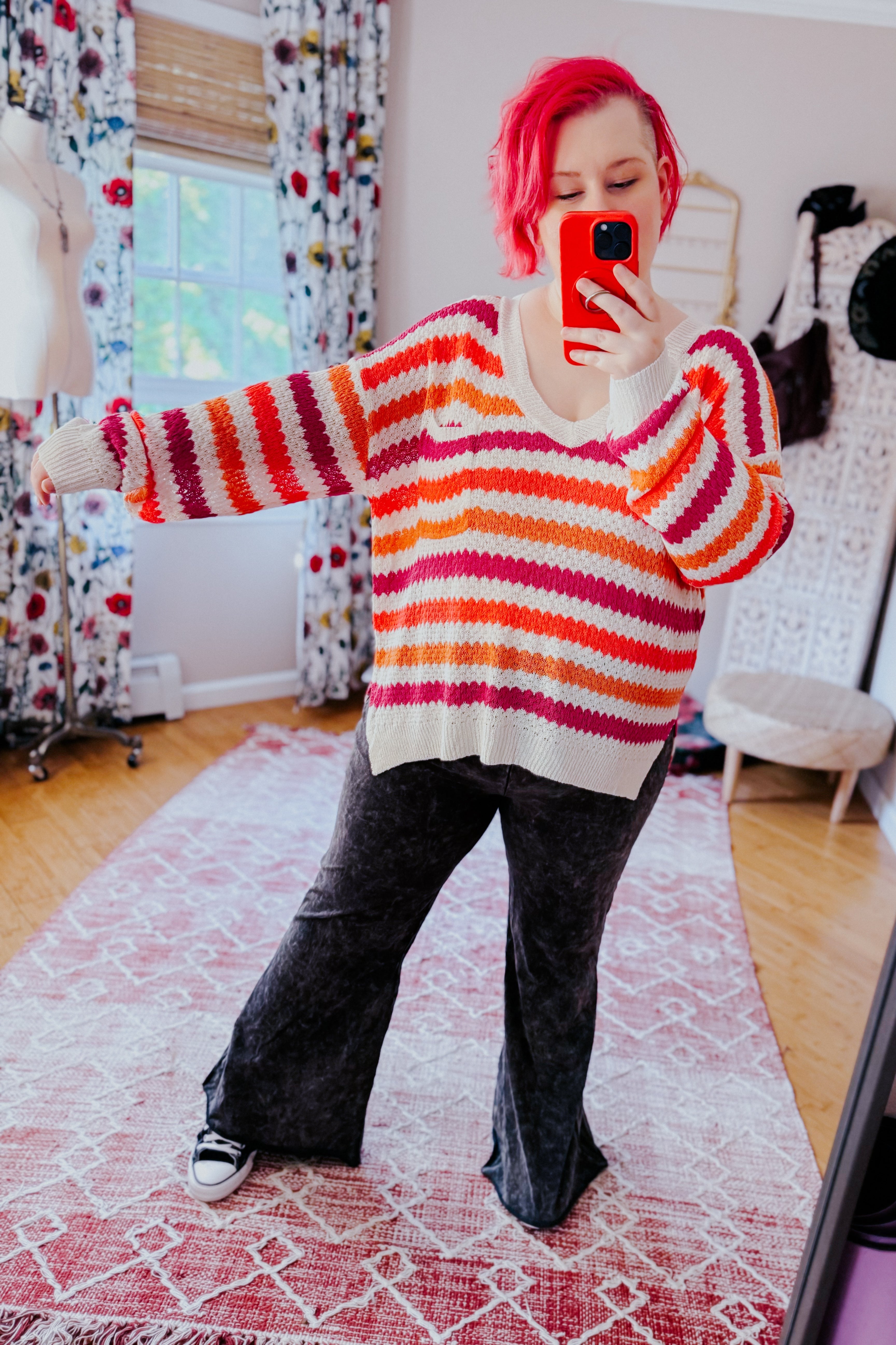 Woman wearing a cozy drop shoulder sweater with pink, orange, and white horizontal stripes paired with loose black pants and sneakers indoors.