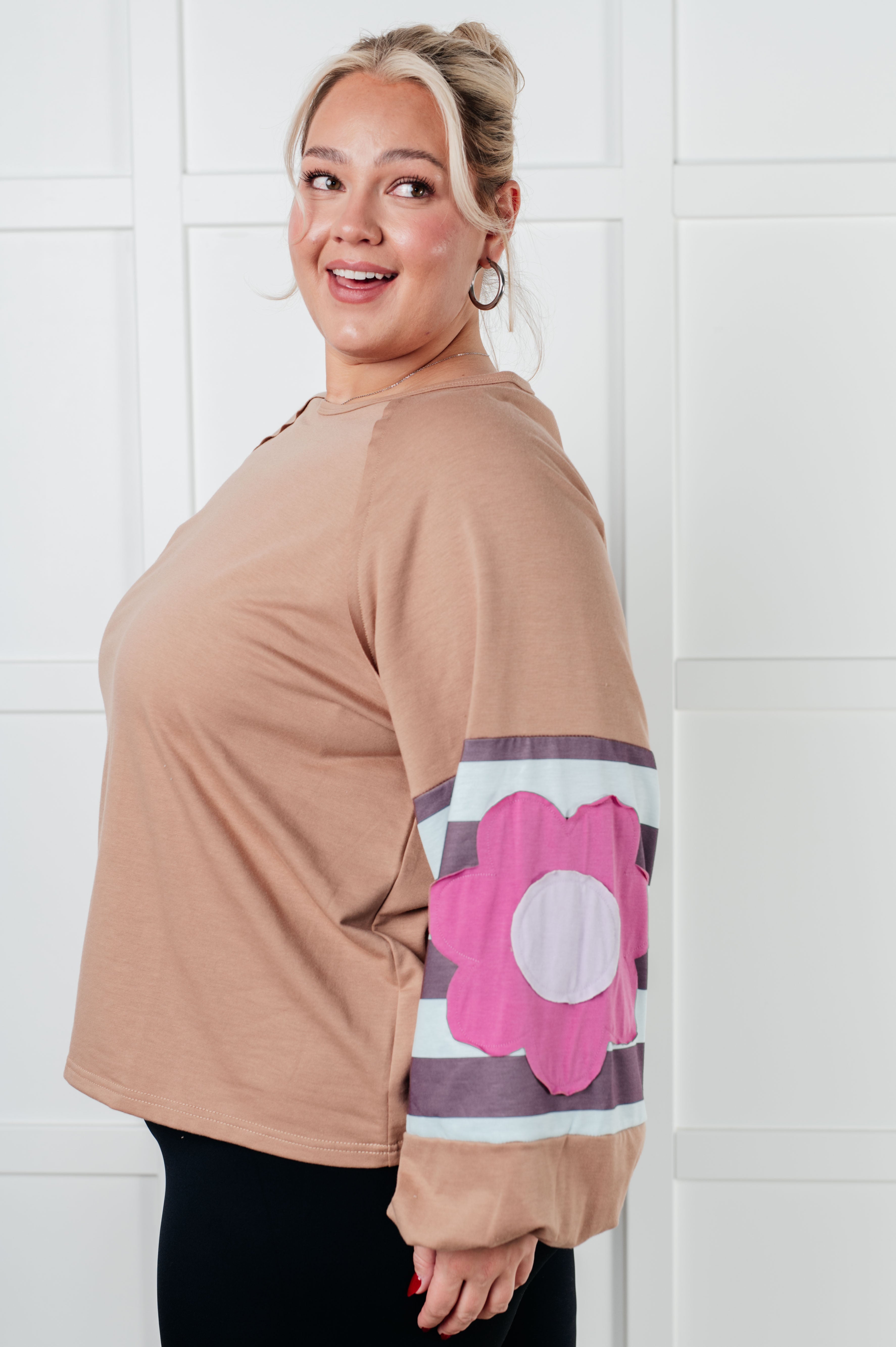 Side view of tan top with striped sleeves and large pink flower patch worn by smiling woman indoors