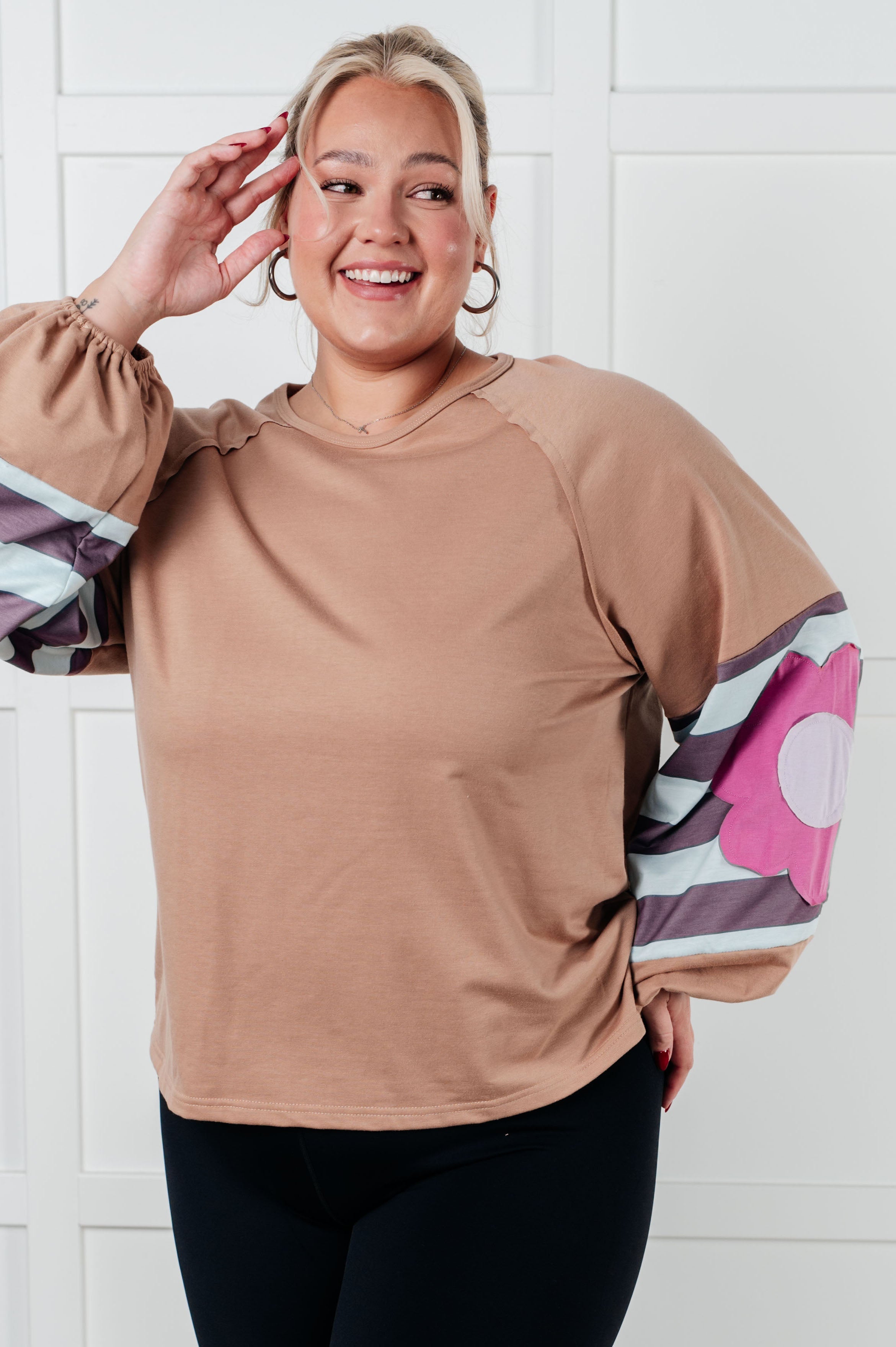 Smiling woman wearing tan pullover with voluminous striped sleeves and large pink floral patch standing indoors