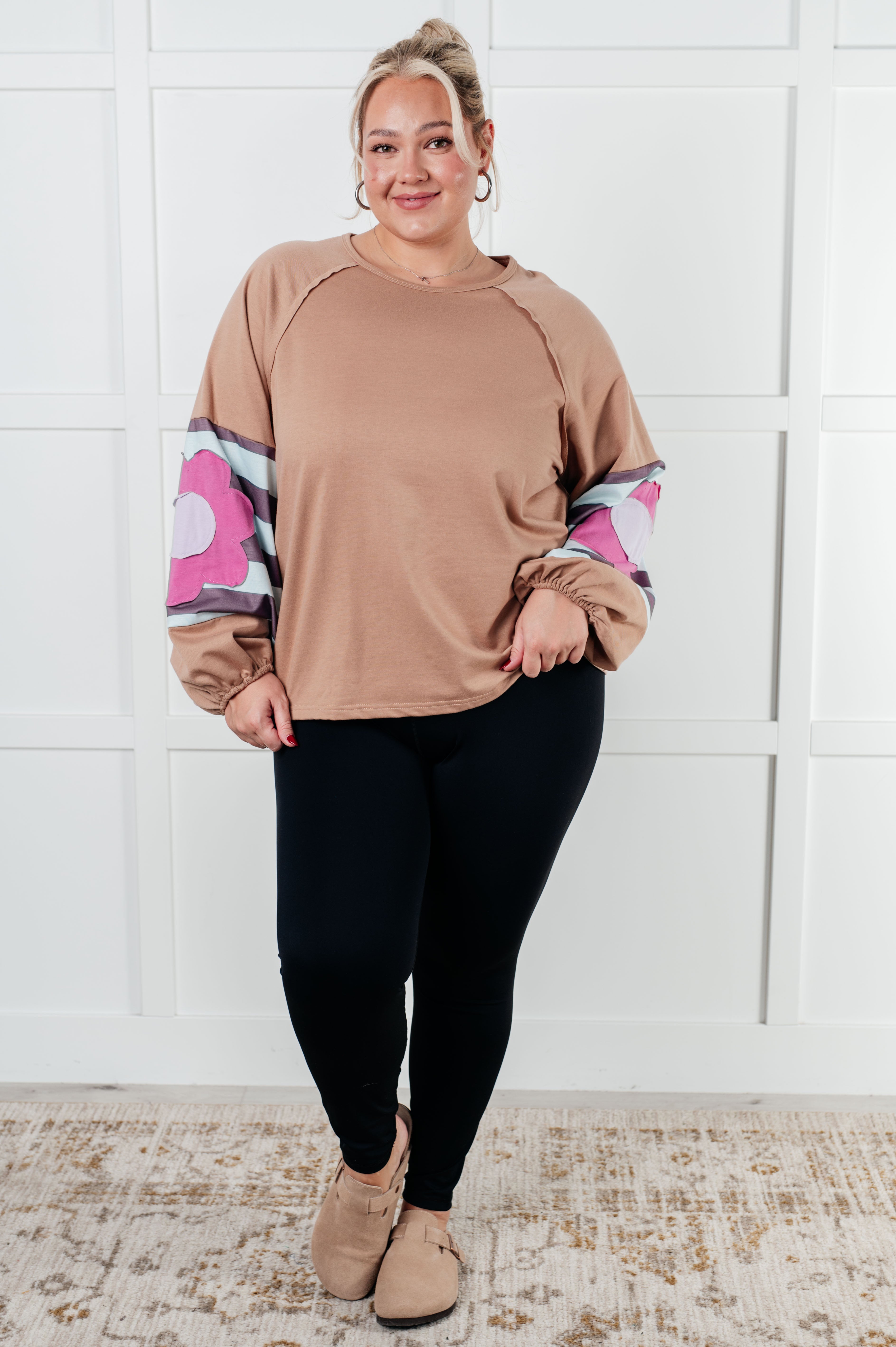 Plus size woman wearing tan floral patchwork top with striped balloon sleeves paired with black leggings and beige shoes indoors