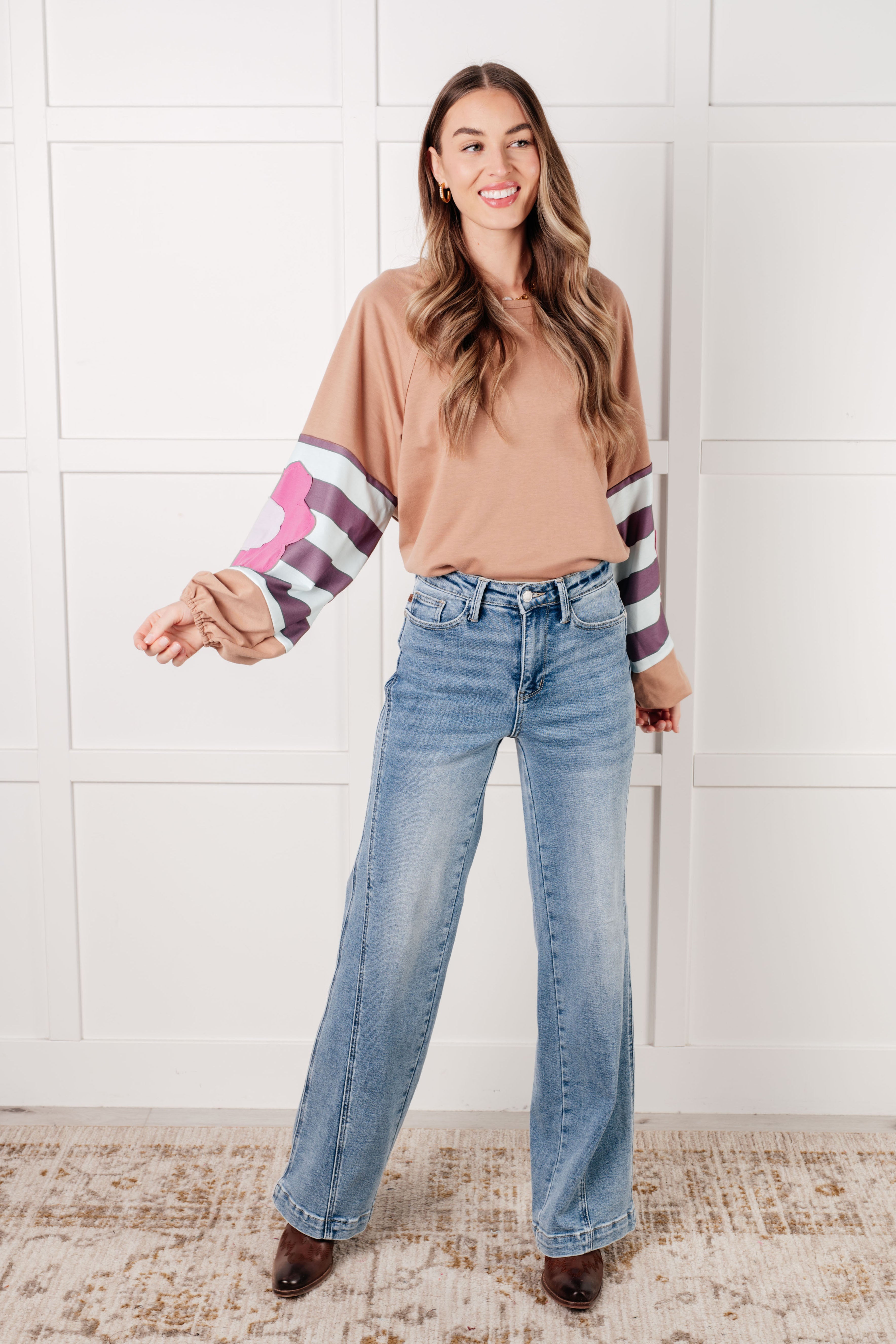 Smiling woman indoors wearing tan top with striped balloon sleeves and large pink floral patch paired with wide-leg blue jeans