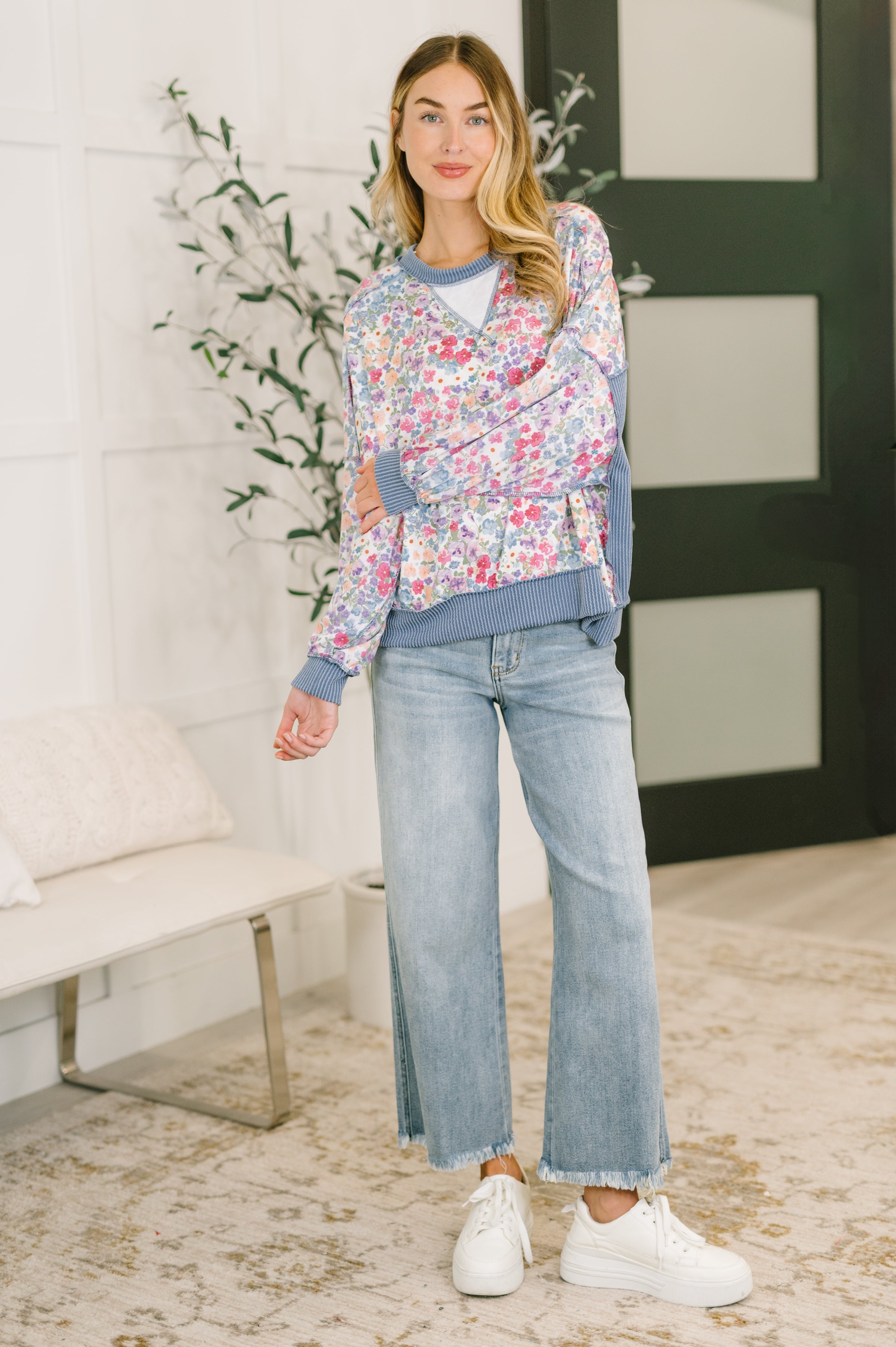 Woman wearing a blue floral drop shoulder pullover with ribbed trim light wash wide leg jeans and white sneakers indoors