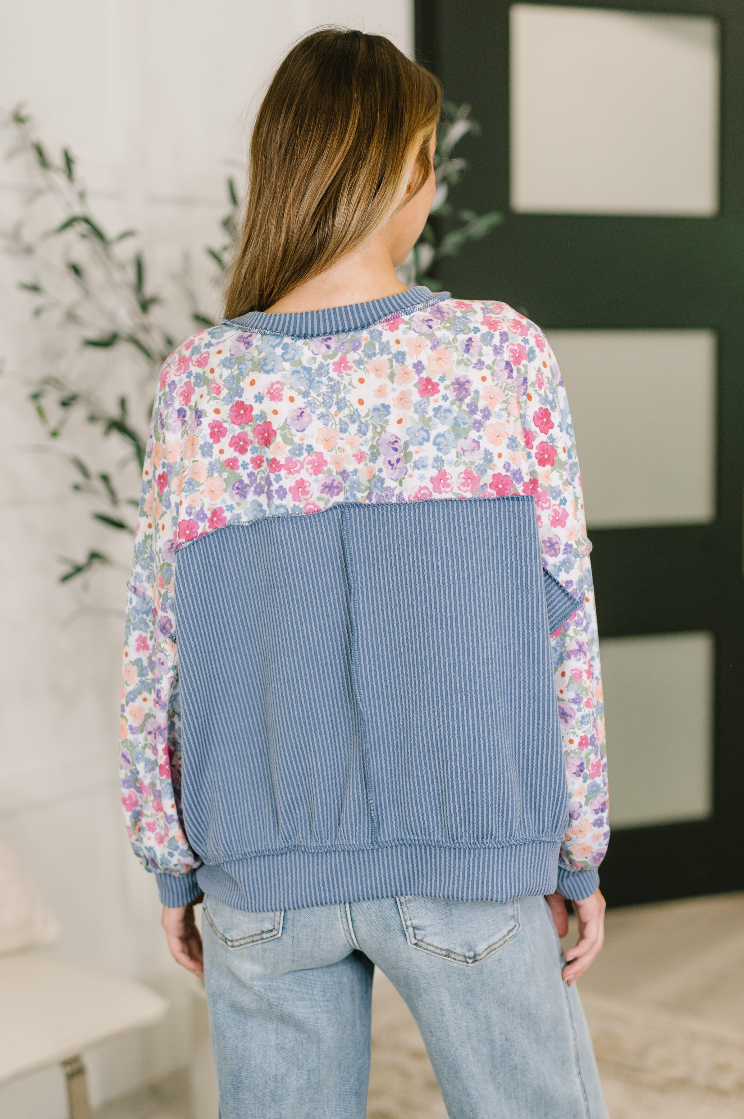 Back view of woman wearing a blue floral pullover with a striped panel and light wash jeans in a softly lit indoor setting