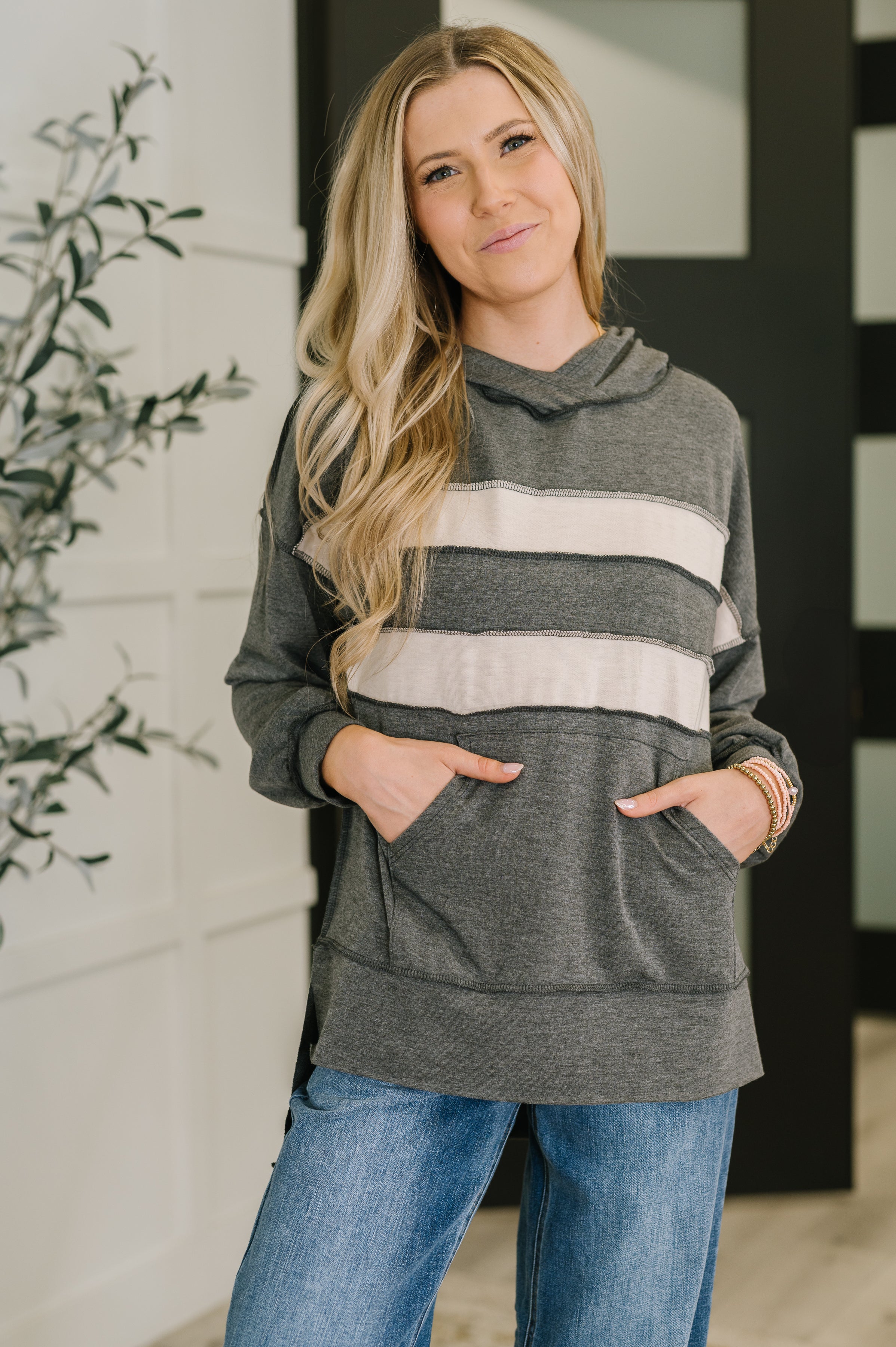 Woman wearing a dark gray hoodie with white horizontal stripes and front pockets, paired with blue jeans, standing indoors near a plant and doors.