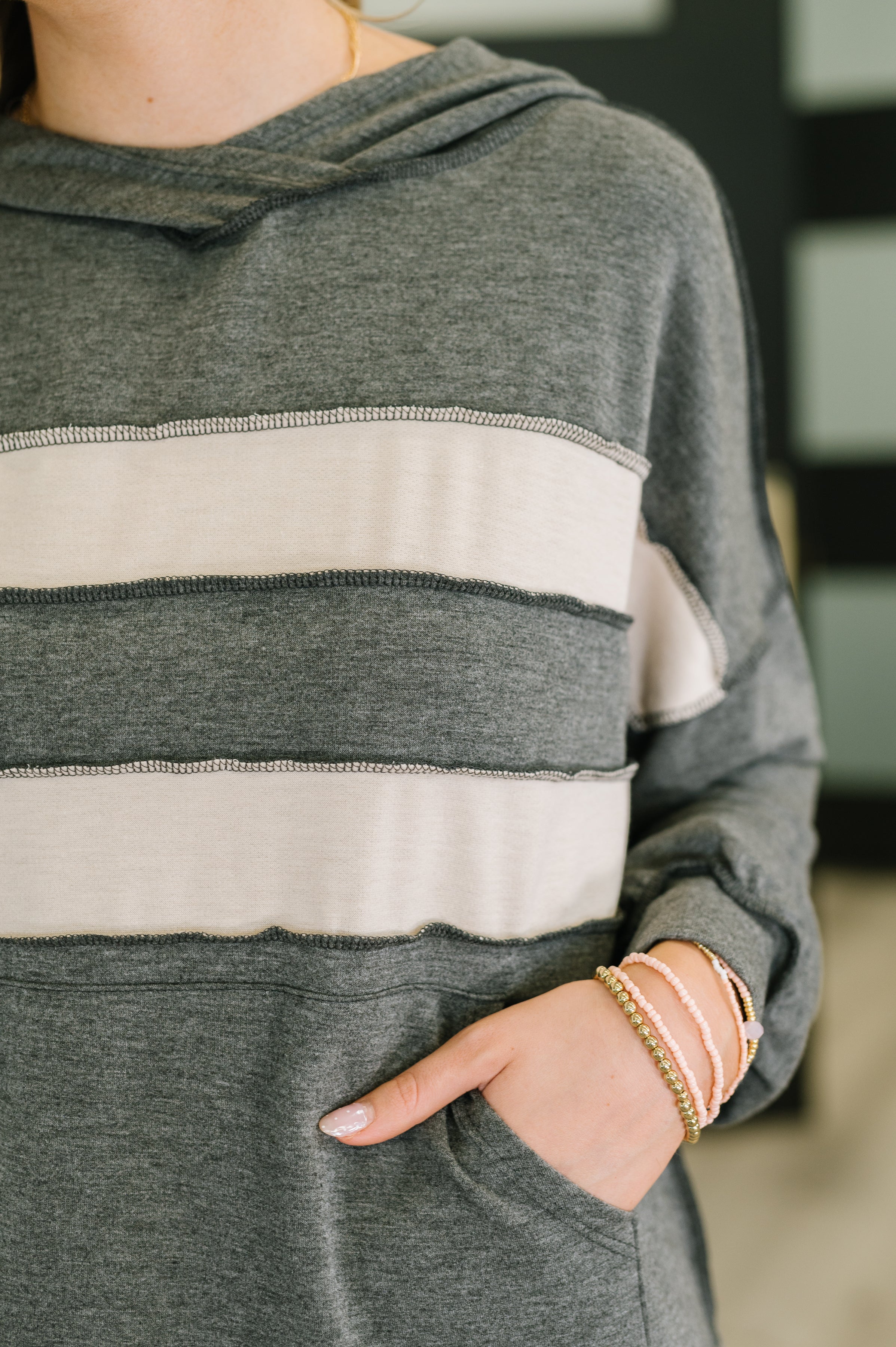 Close-up of a person wearing a gray pullover with wide cream horizontal stripes and raw-edge seams, their hand resting in a front pocket and adorned with beaded bracelets.