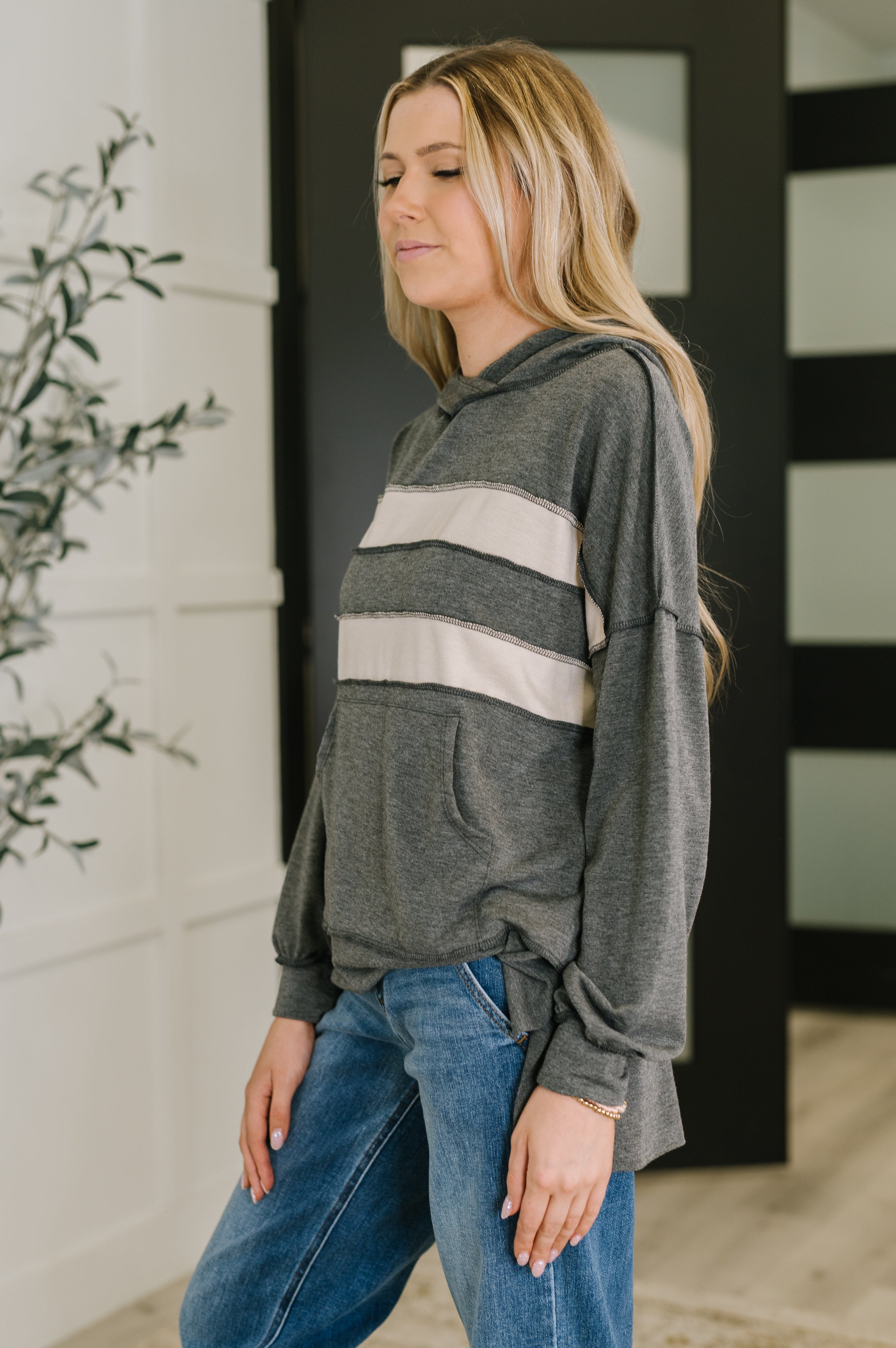 Side view of a woman wearing a gray pullover hoodie with two wide cream horizontal stripes, paired with relaxed blue jeans indoors near a plant and modern doors.