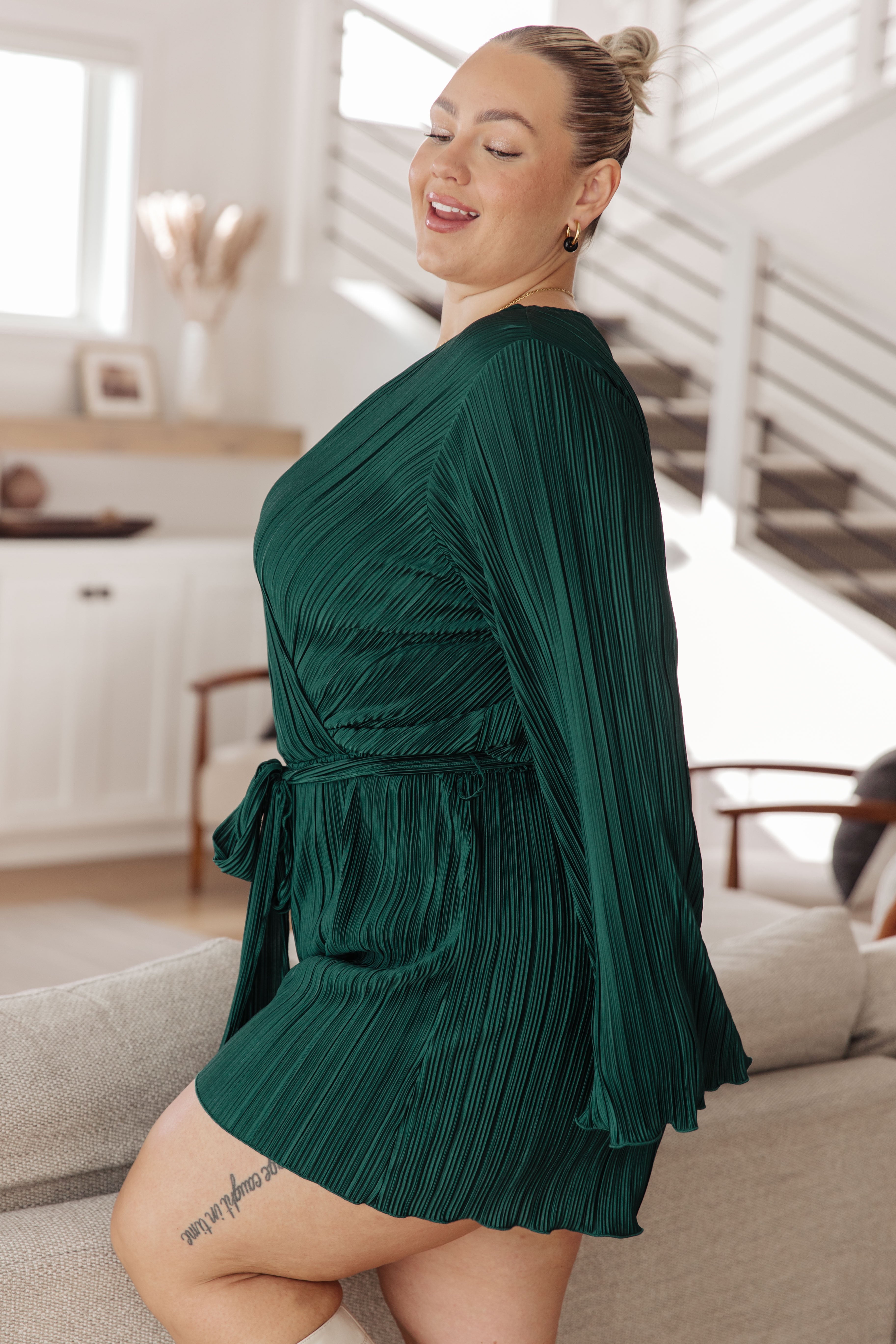Plus-size woman indoors smiling while wearing a hunter green pleated romper with long flowing sleeves and a tie waist, seated on a beige couch.