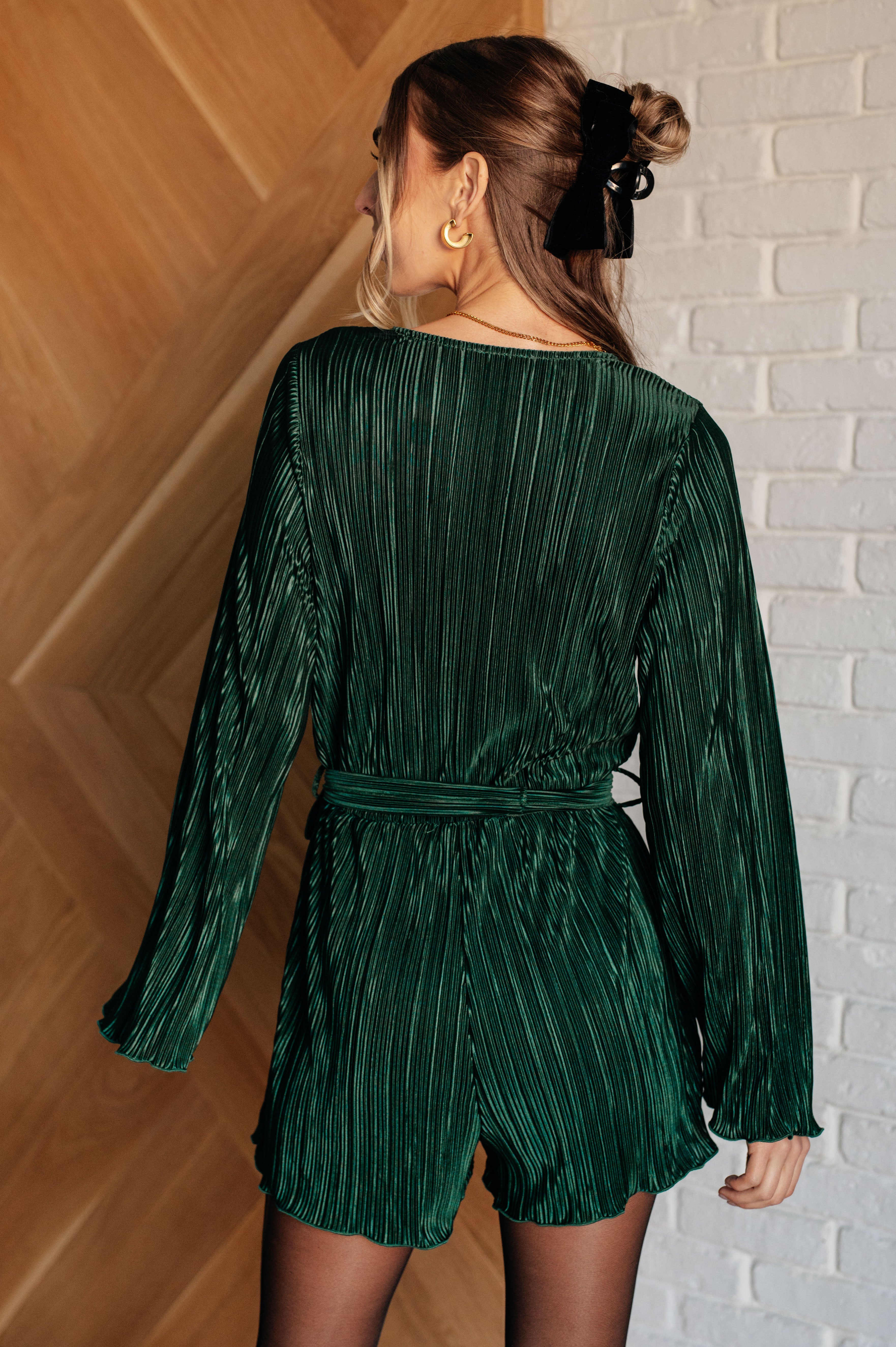 Woman shown from behind wearing a hunter green pleated romper with long sleeves and a waist tie, styled with a black hair accessory indoors.