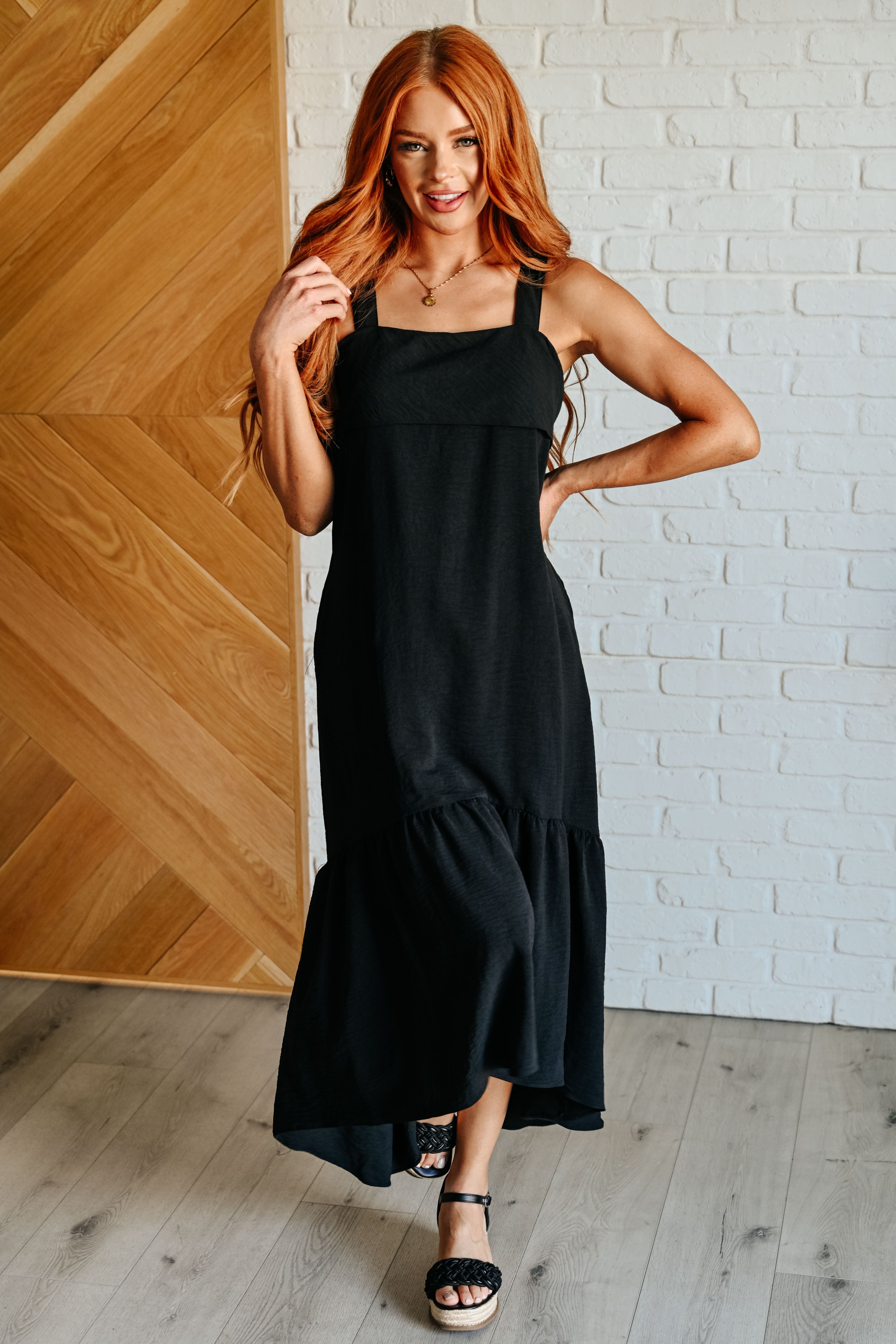 Woman wearing black maxi dress with square neckline and wide straps paired with black platform sandals indoors