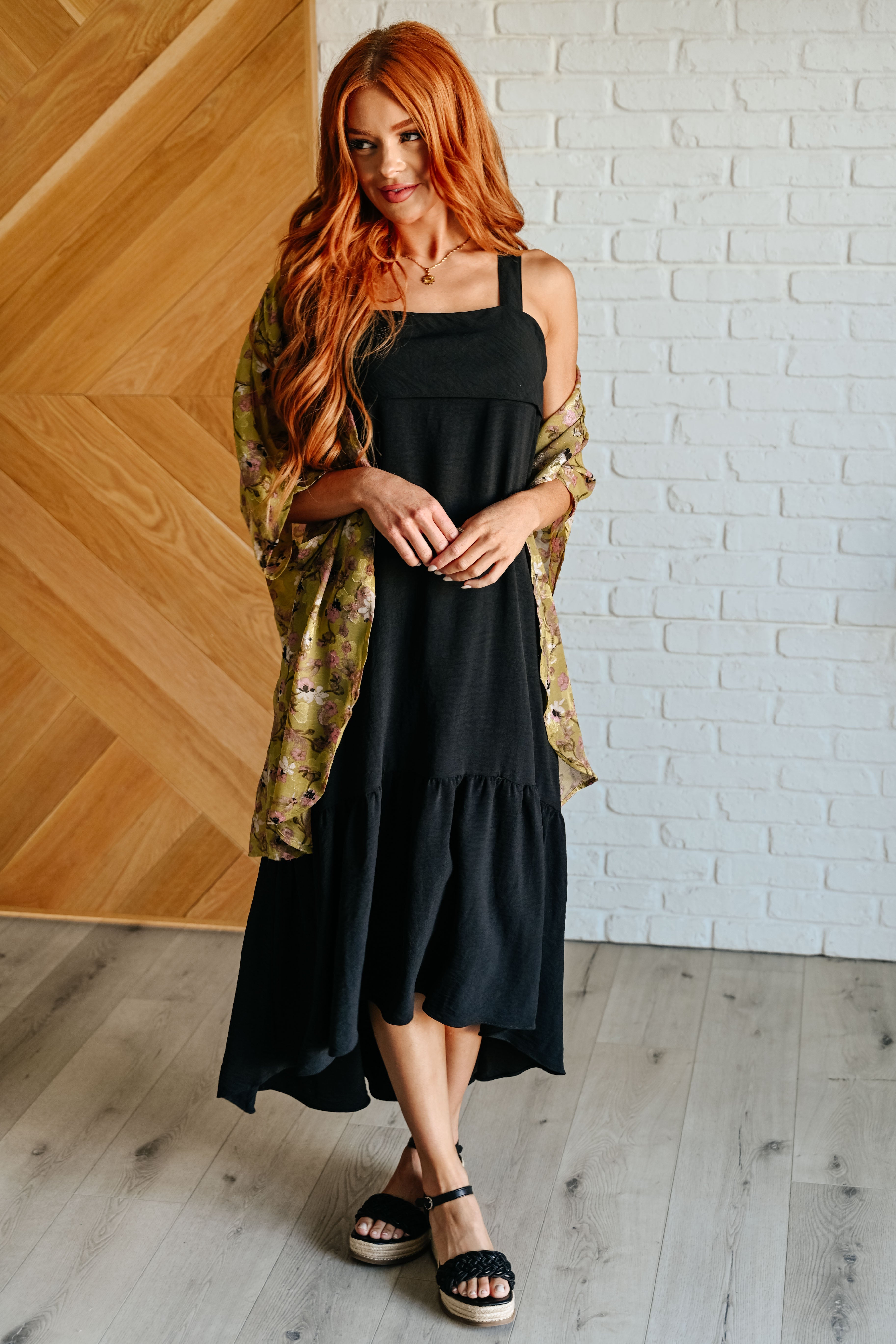 Woman with long red hair wearing a black sleeveless maxi dress layered with a green floral shawl and black platform sandals