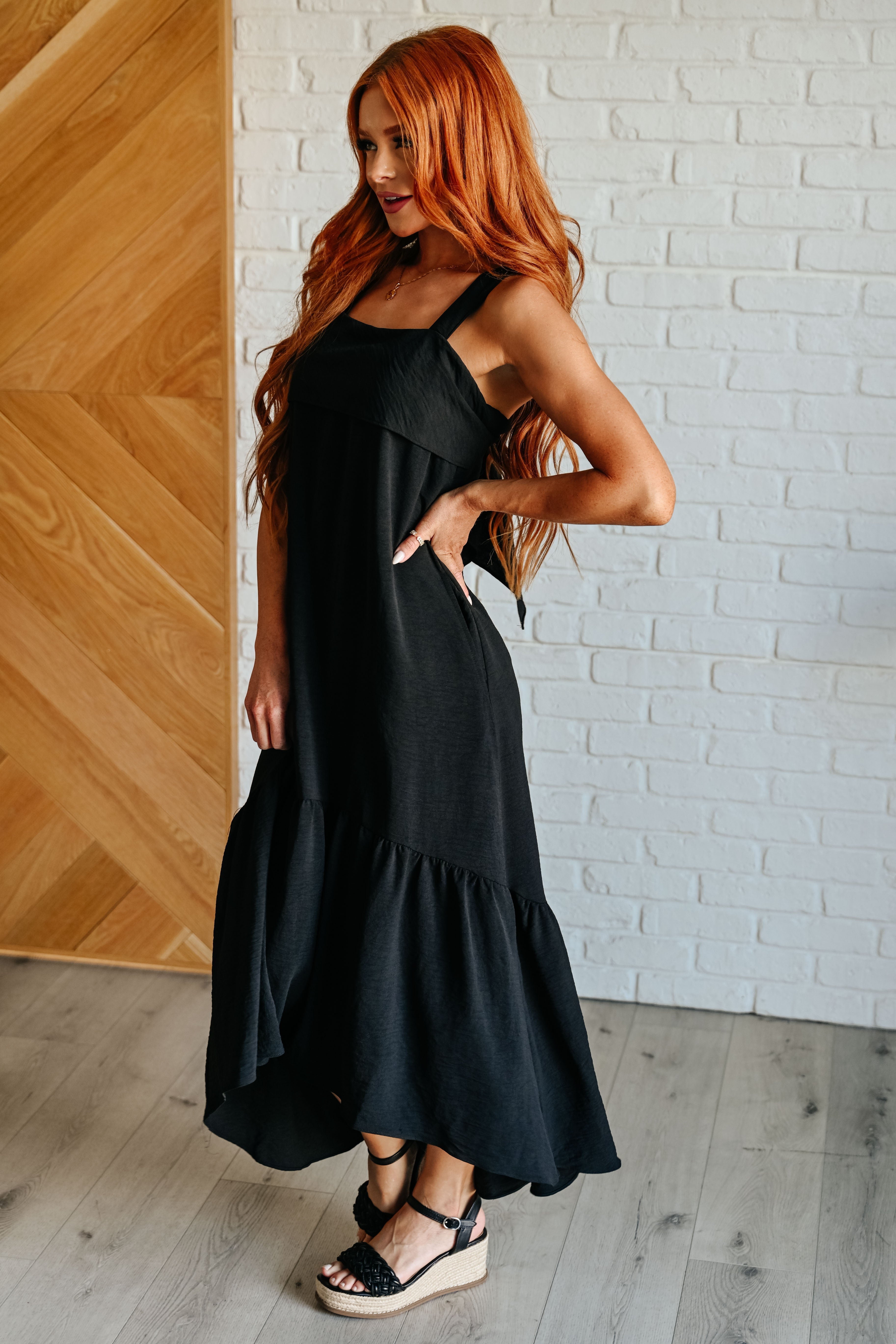 Woman with long red hair wearing black tie back maxi dress and black wedge sandals against white brick wall