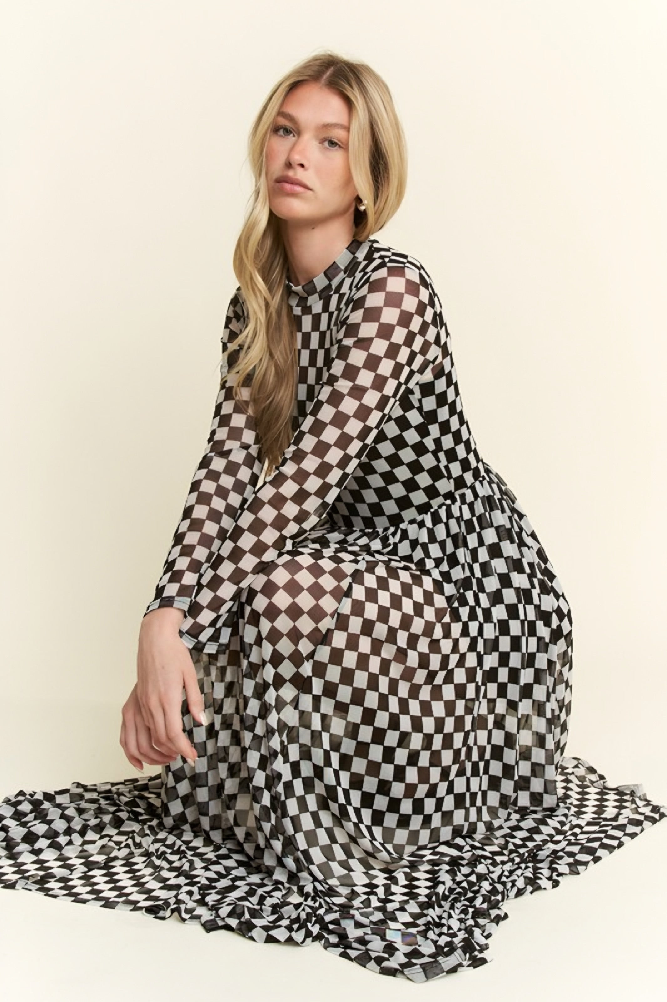 Woman seated wearing a black and white checkered print mesh maxi dress with long sleeves and a high neckline against a light background.