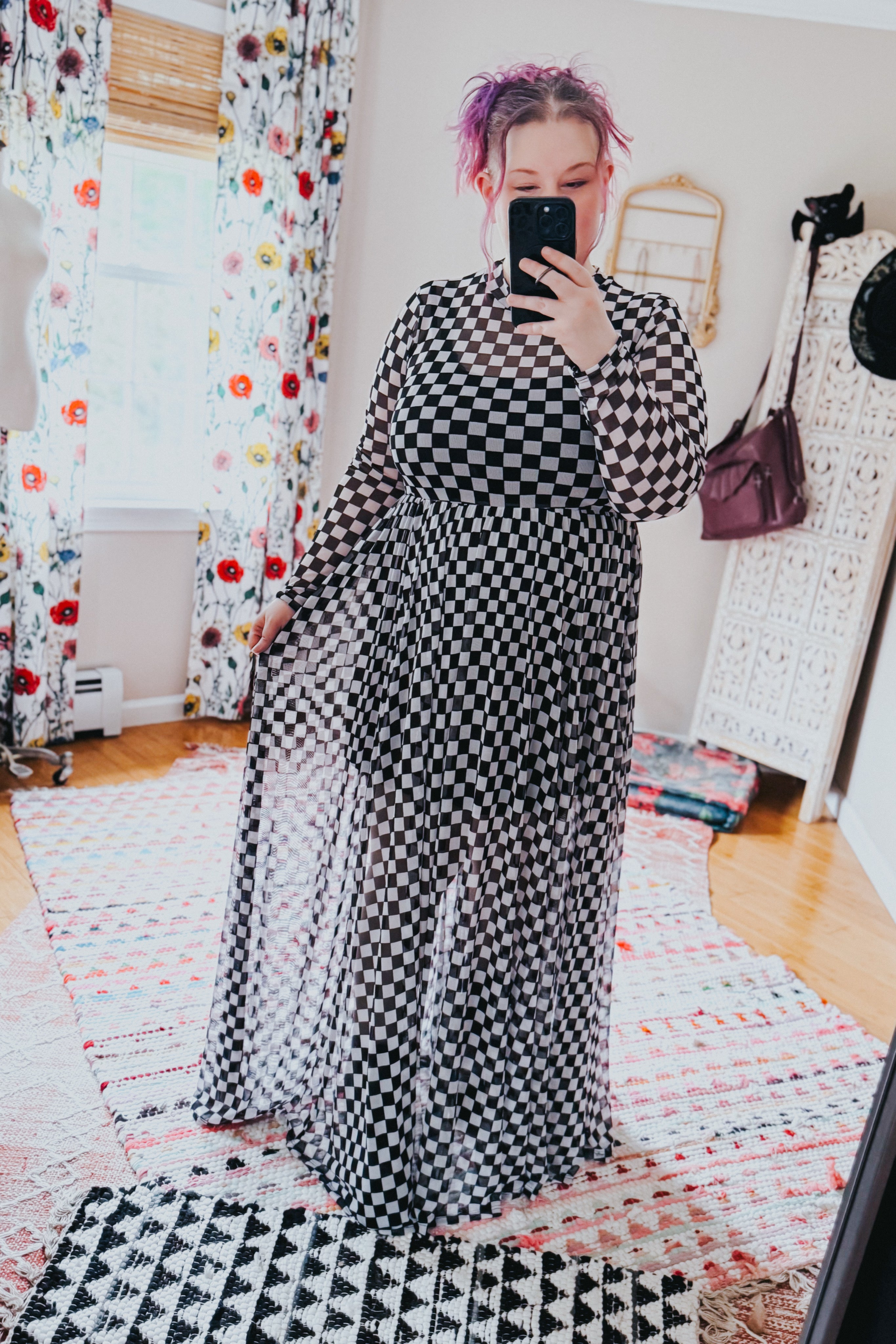 Woman taking a mirror selfie wearing a black and white checkered print long-sleeve mesh maxi dress while holding the skirt in a bright room.