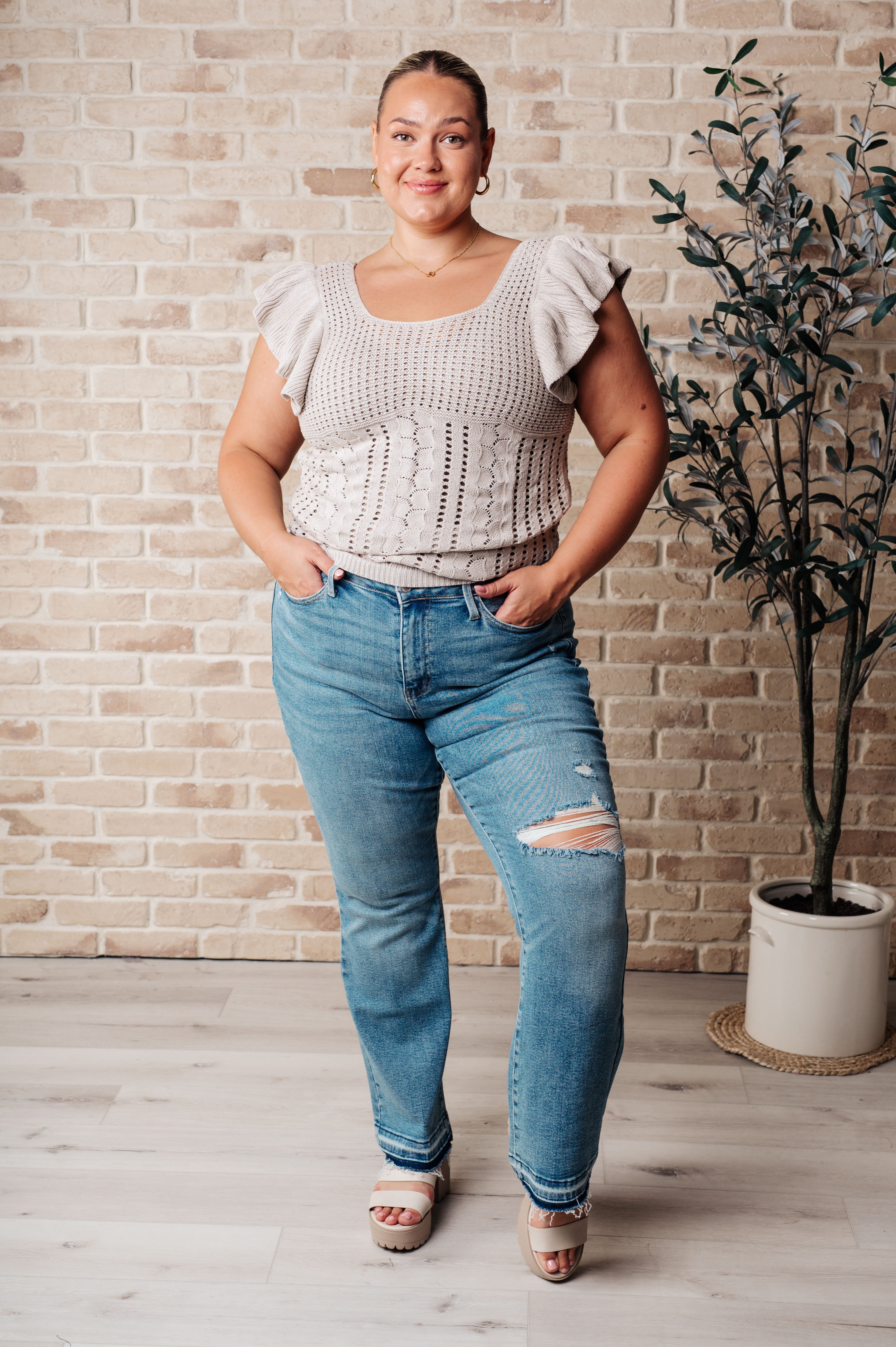 Confident woman wearing a grey pointelle knit top with ruffle sleeves paired with distressed blue jeans and white platform sandals against a brick wall