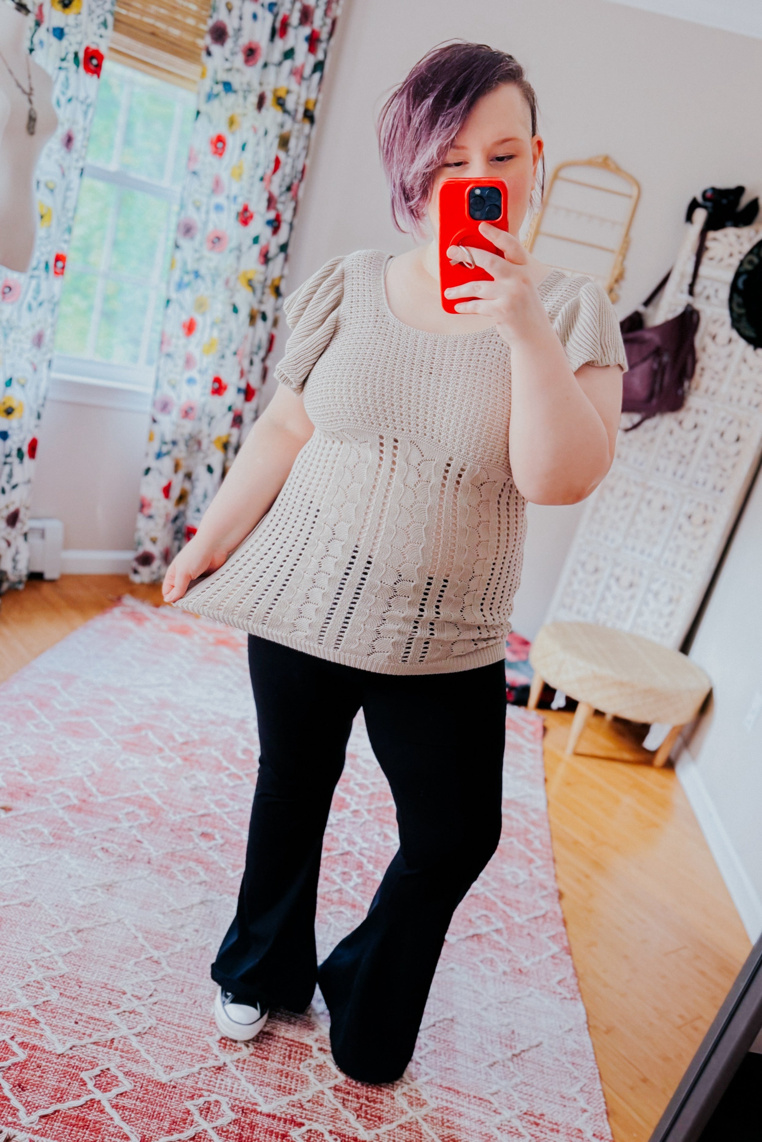 Woman with short purple hair taking a mirror selfie wearing a grey knit top with puffed sleeves paired with black flared pants in a bright room with floral curtains and a patterned rug