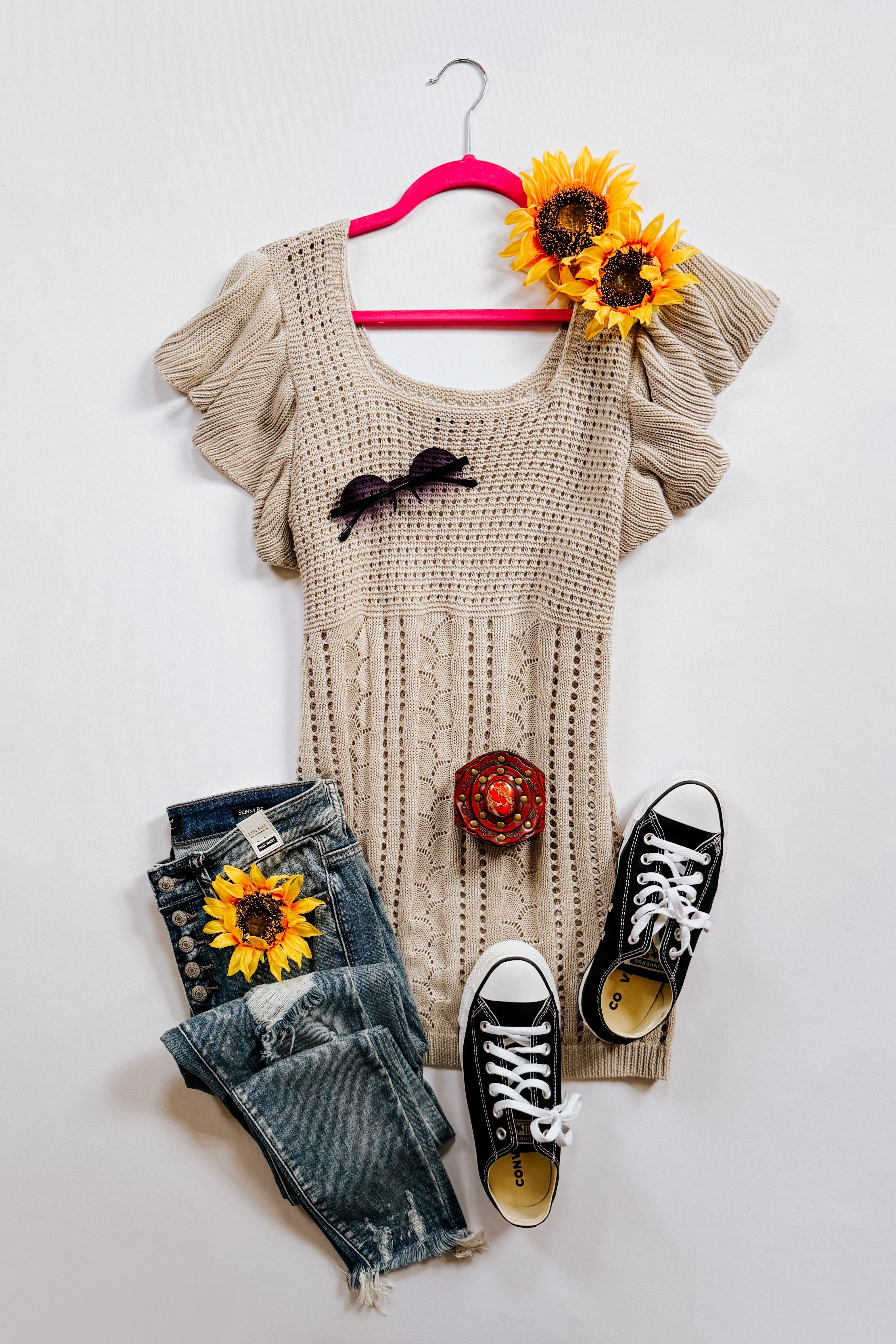 Grey pointelle knit top with ruffled short sleeves displayed on a hanger alongside distressed blue jeans, black sneakers, and sunflower accessories.