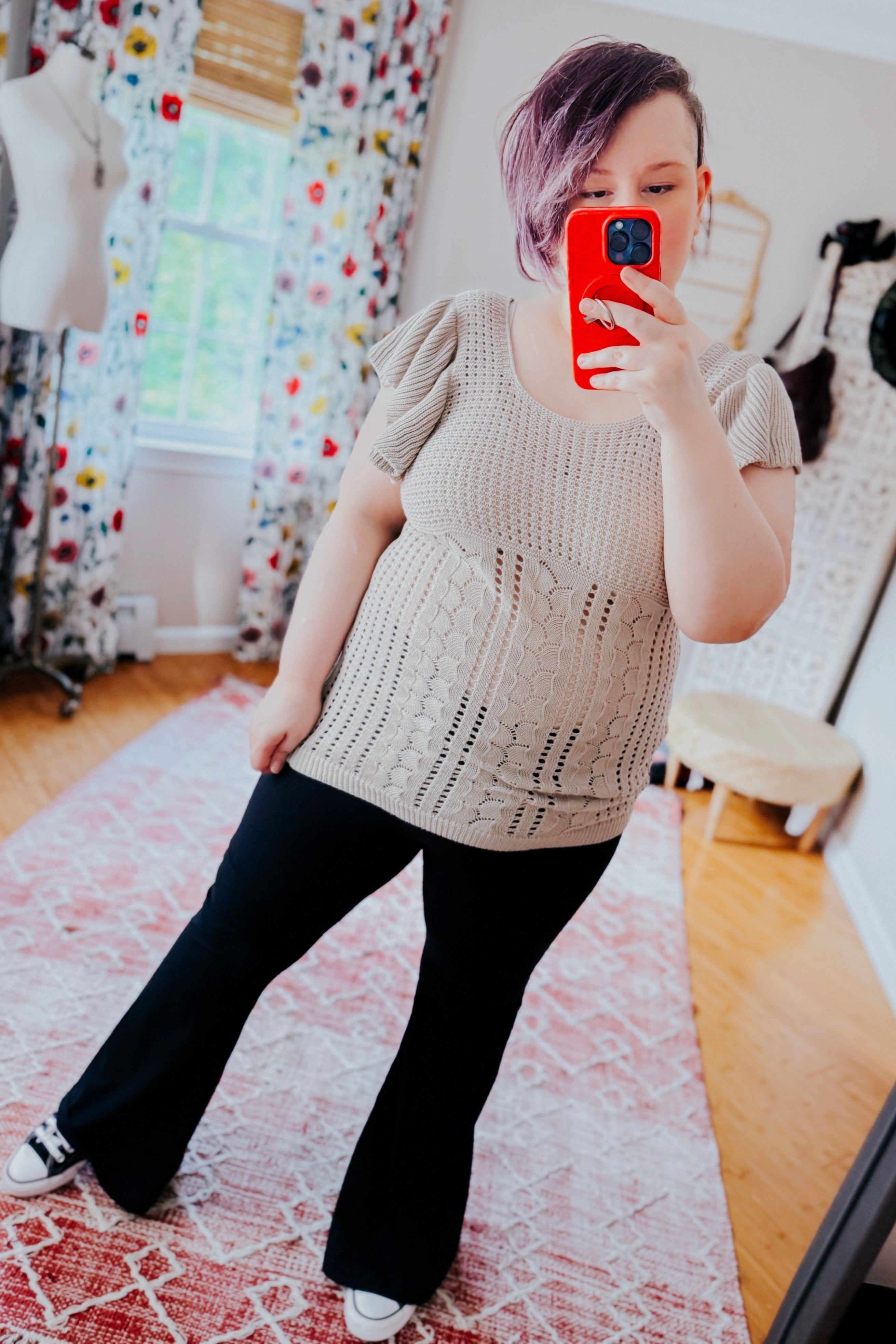 Woman taking a mirror selfie wearing a grey pointelle knit top with puffed sleeves and black flared pants in a bright room with floral curtains and a red patterned rug.