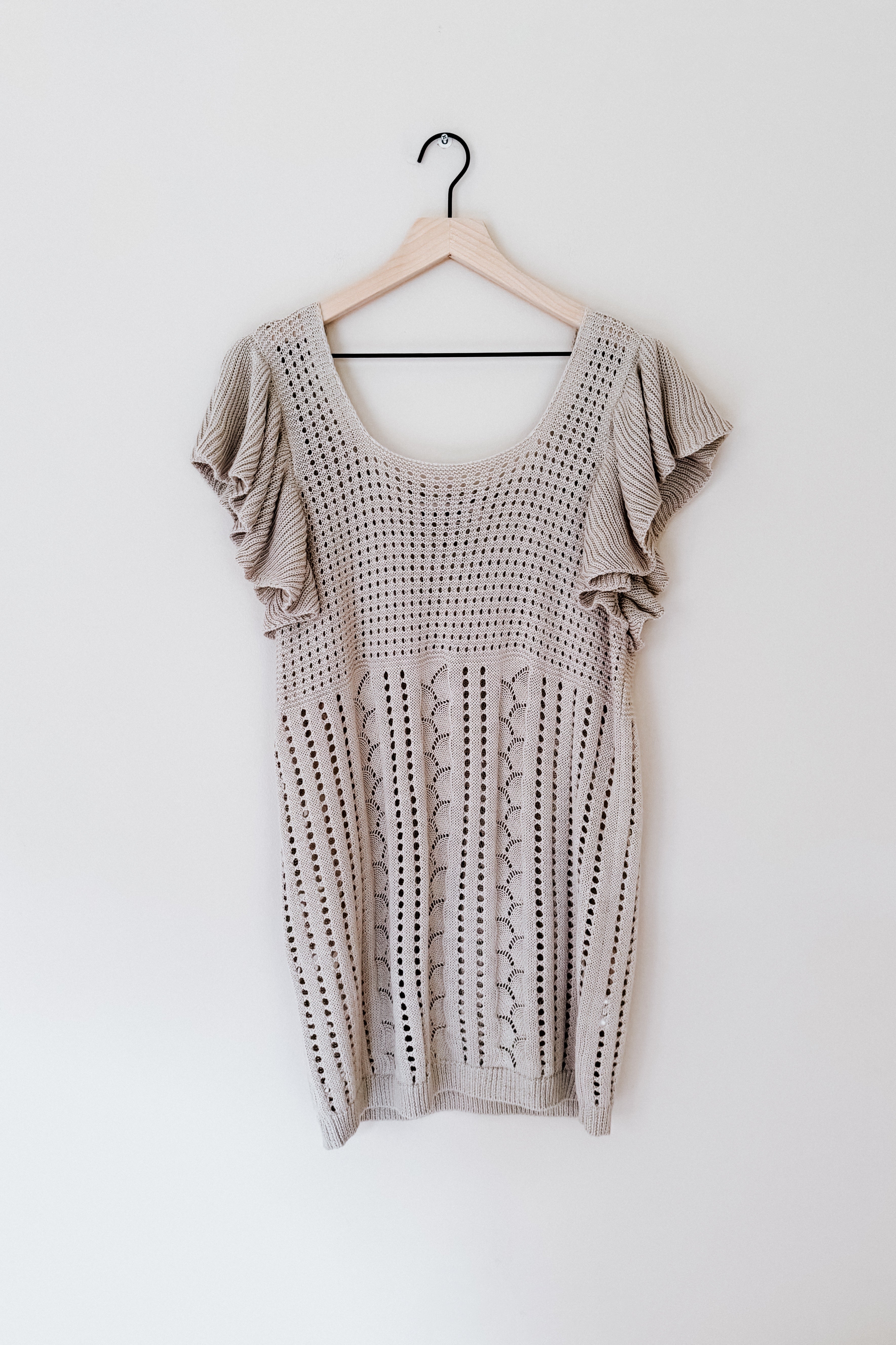 Light grey knit top with puffed short sleeves and intricate pointelle pattern hanging on a wooden hanger against a plain white wall.
