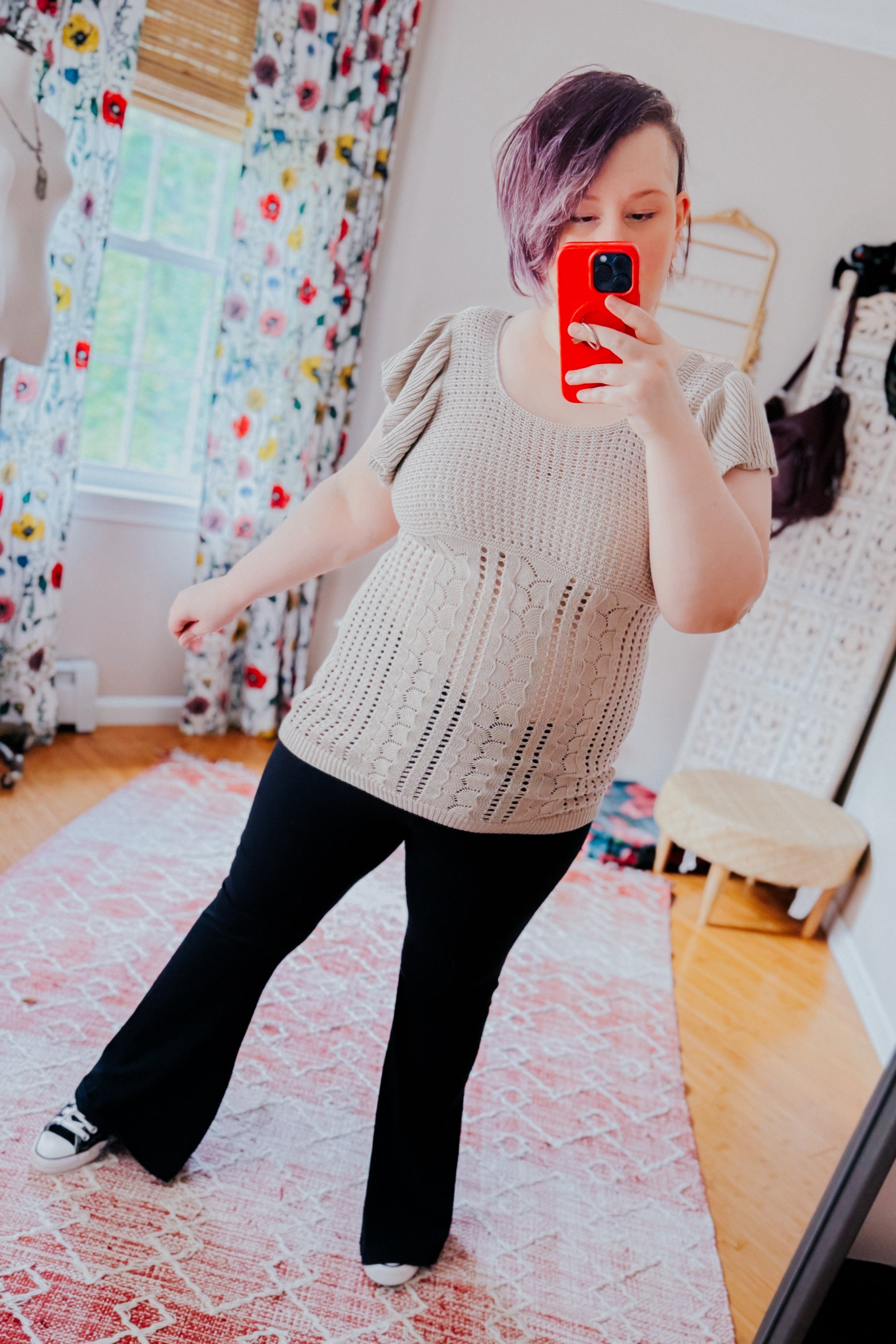 Woman with short purple hair wearing a grey textured knit top with flutter sleeves and black pants, taking a mirror selfie in a bright room with floral curtains and a pink patterned rug.