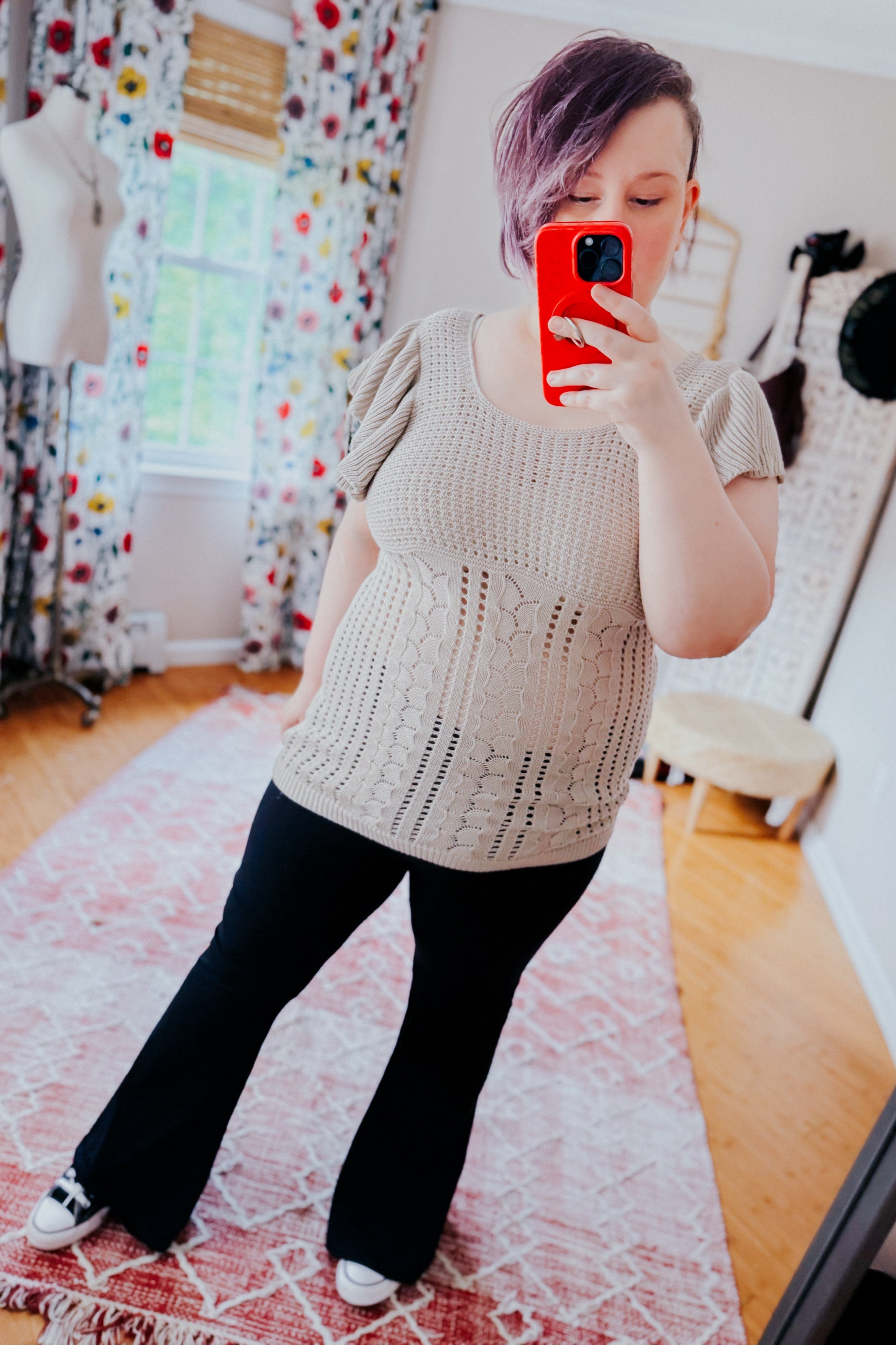 Woman with short purple hair wearing a grey pointelle knit top with puffed sleeves and black pants, taking a mirror selfie in a bright room with floral curtains and a pink patterned rug.
