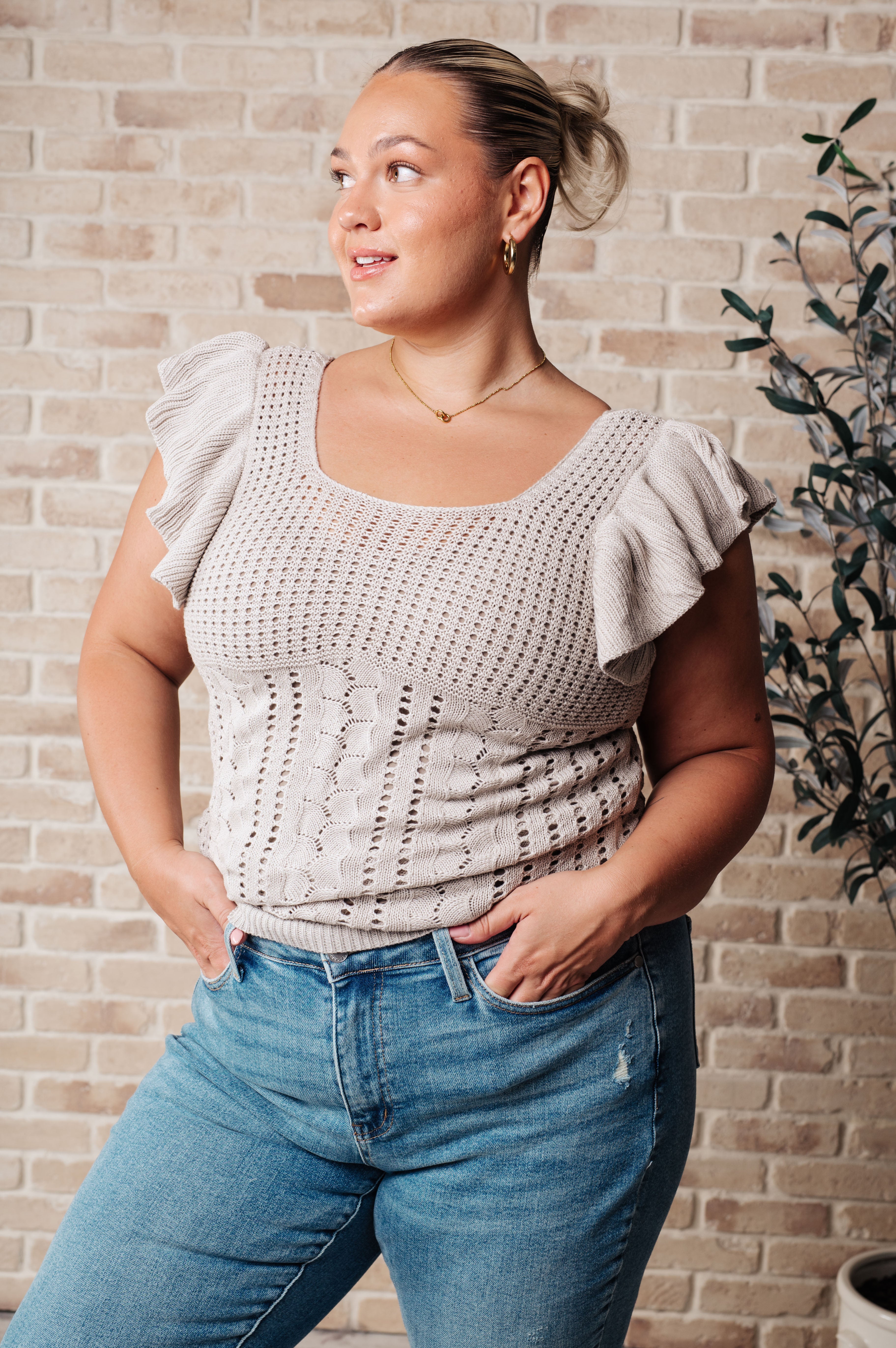 Curvy woman wearing a grey pointelle knit top with flutter sleeves paired with light blue distressed jeans, standing against a brick wall.