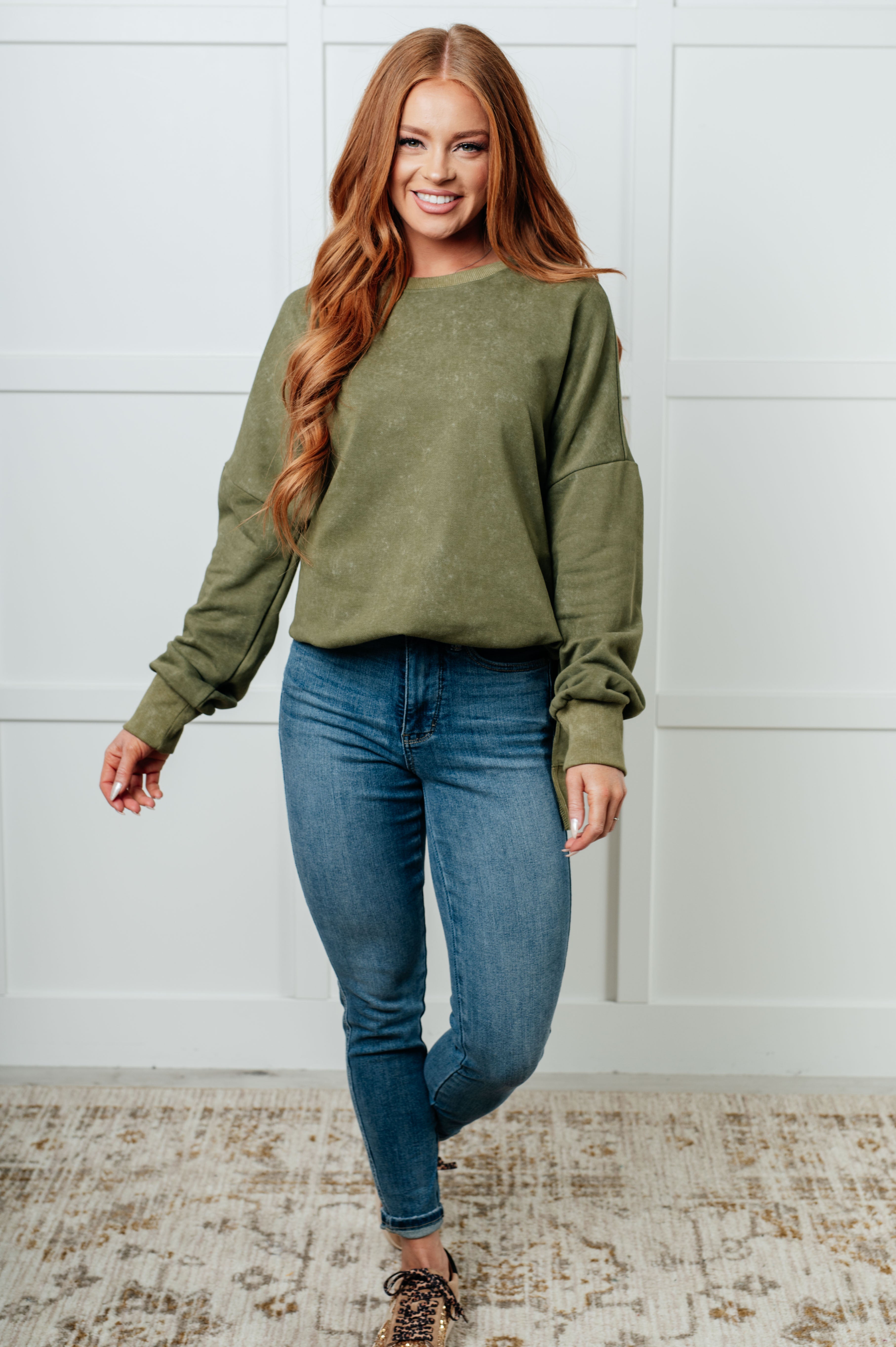Smiling woman with long red hair wearing a green oversized sweatshirt paired with fitted blue jeans and leopard-print sneakers, standing indoors.