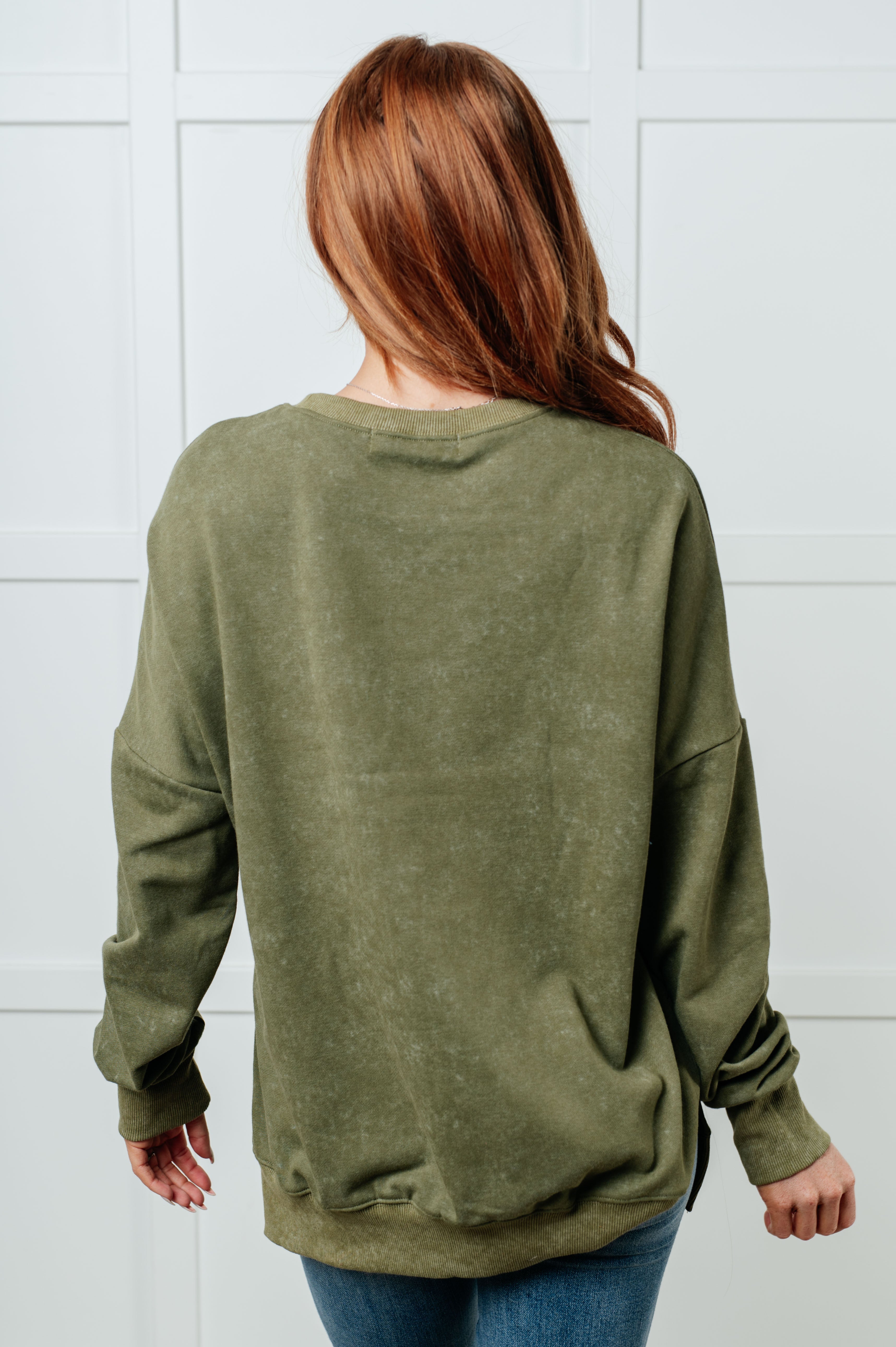 Woman with long red hair modeling a relaxed olive green oversized sweatshirt from the back against a white paneled wall background.