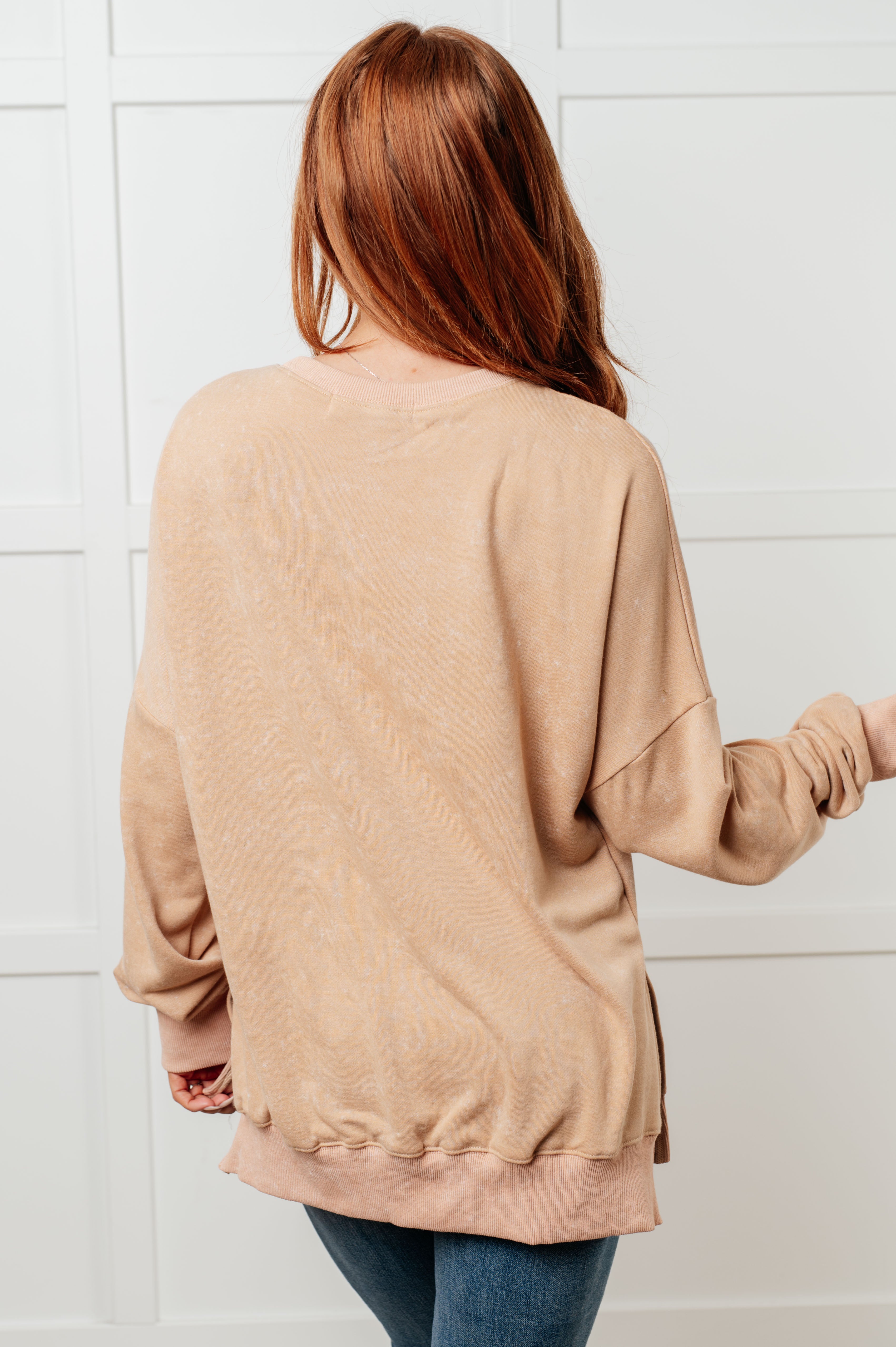 Woman with long red hair wearing a loose-fitting khaki sweatshirt with ribbed hem and cuffs, shown from the back against a white paneled wall.