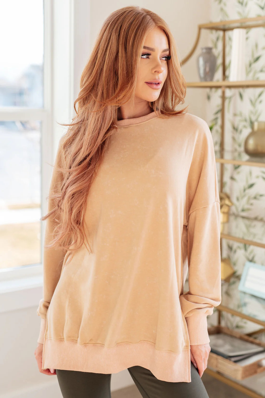 Woman with long red hair wearing a soft khaki oversized sweatshirt with relaxed fit and ribbed cuffs, standing indoors near a window and shelf
